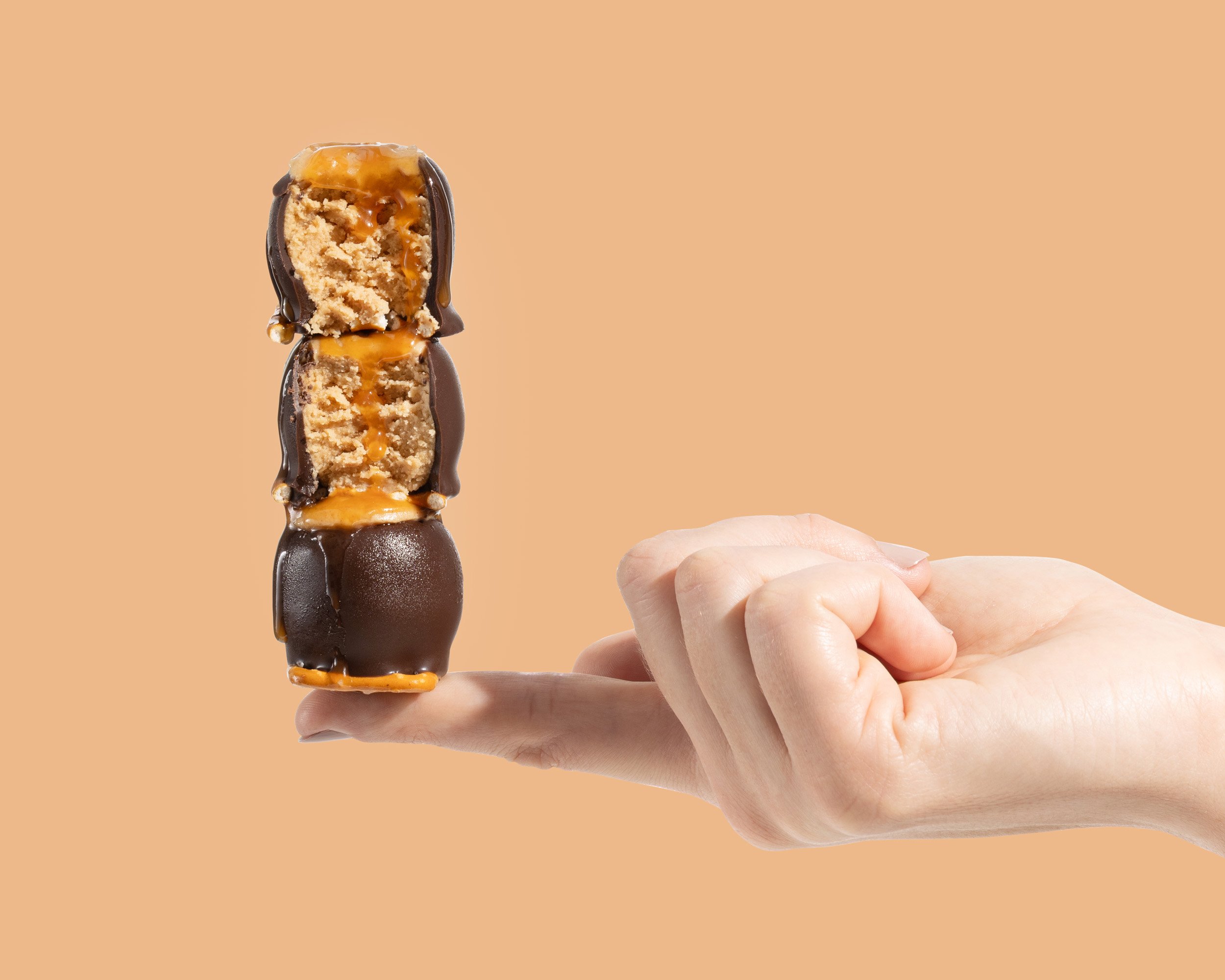 A hand holding a stack of three caramel-filled cream puff pastries dipped in chocolate, against a peach-colored background.