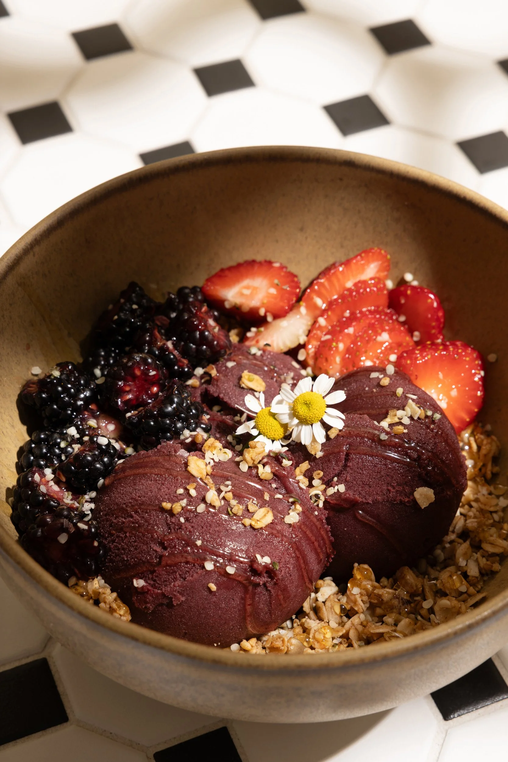A bowl of frozen chocolate ice cream topped with chopped nuts, fresh strawberries, blackberries, and edible flowers, on a black and white checkered surface.