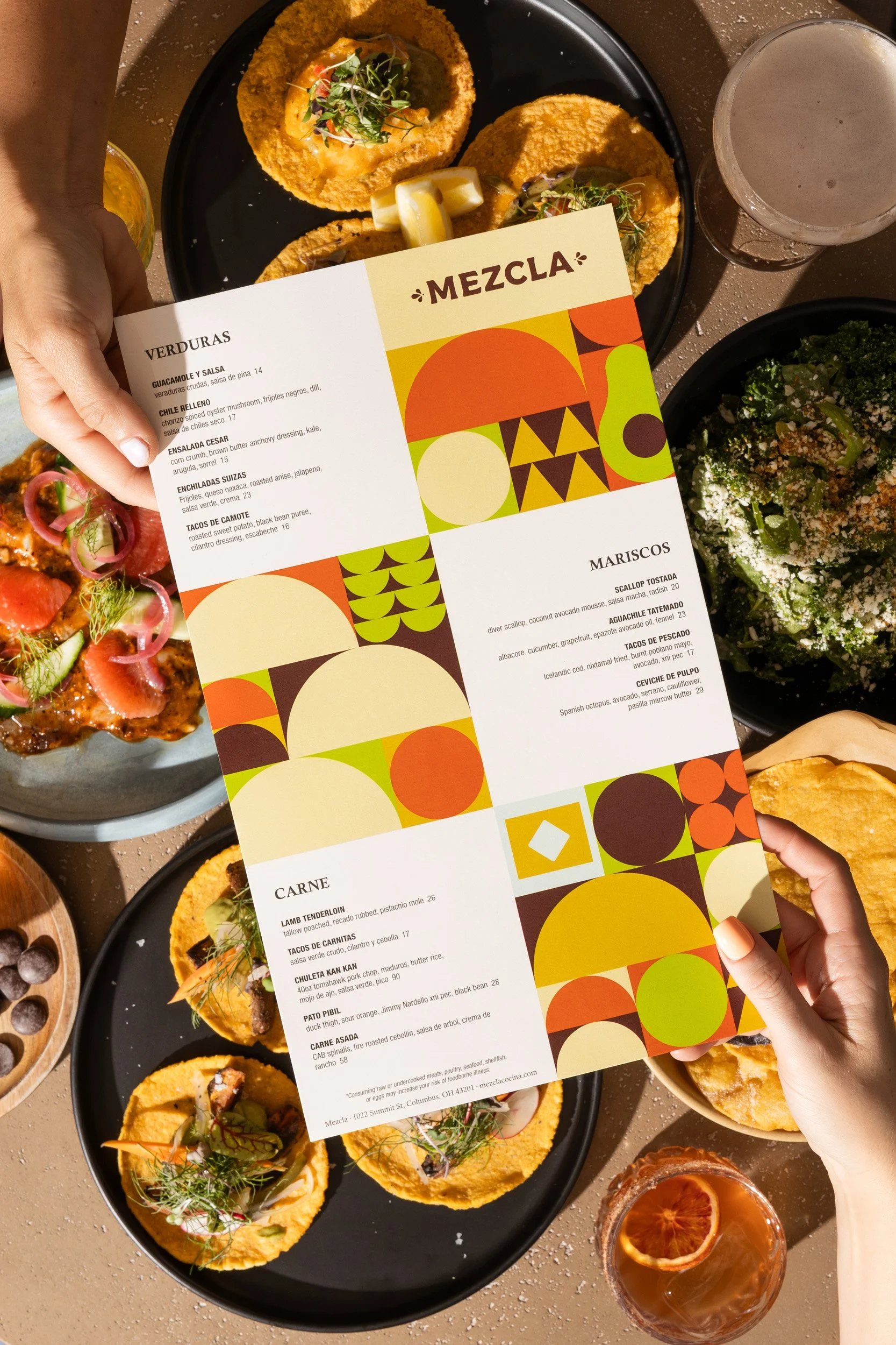 Person holding a colorful menu over a table of Mexican dishes, including tacos, ceviche, a salad, and drinks.