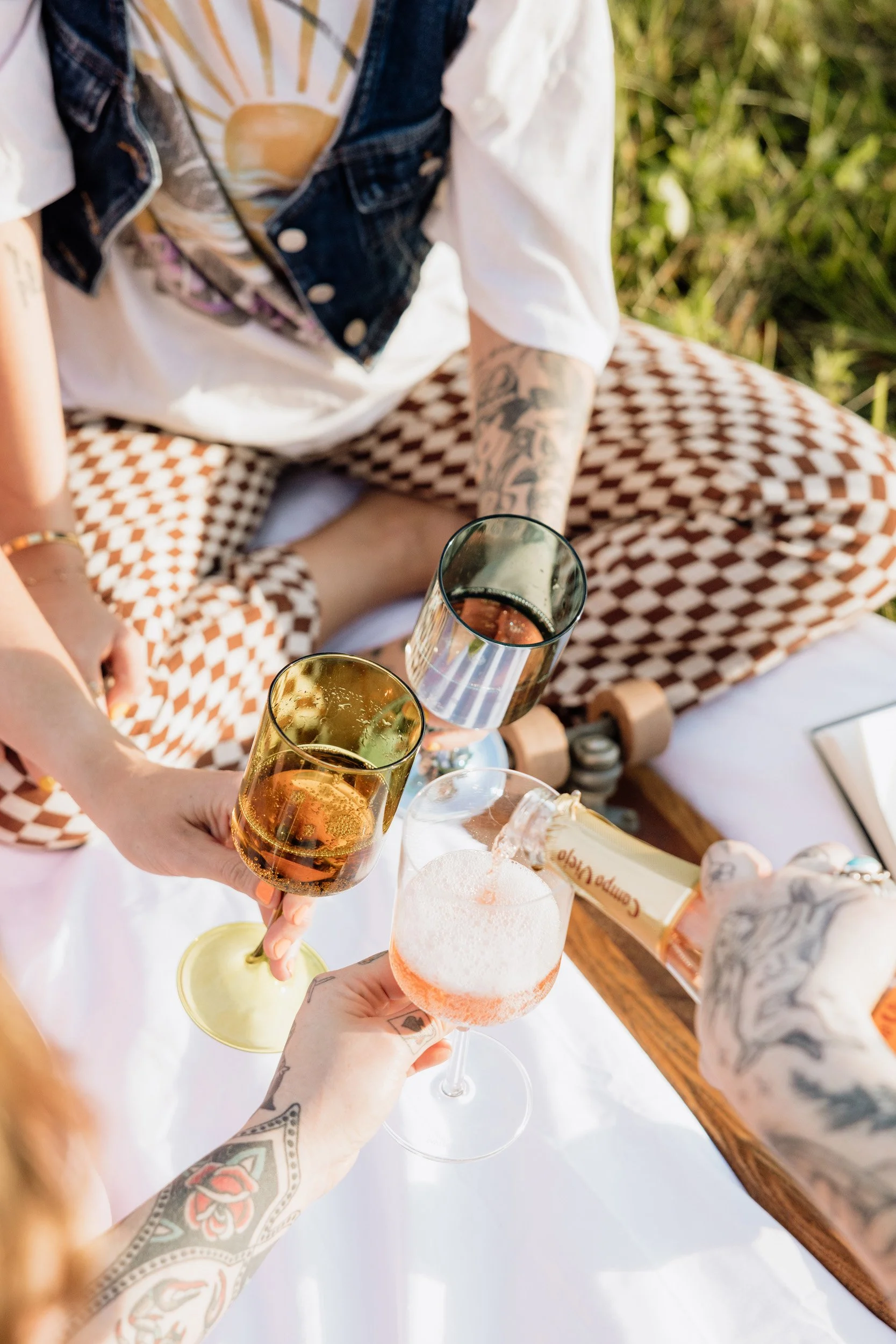 People gathered outdoors on a white table, raising glasses with drinks such as champagne, rosé, and soda in a toast. One person is pouring a drink into a glass.