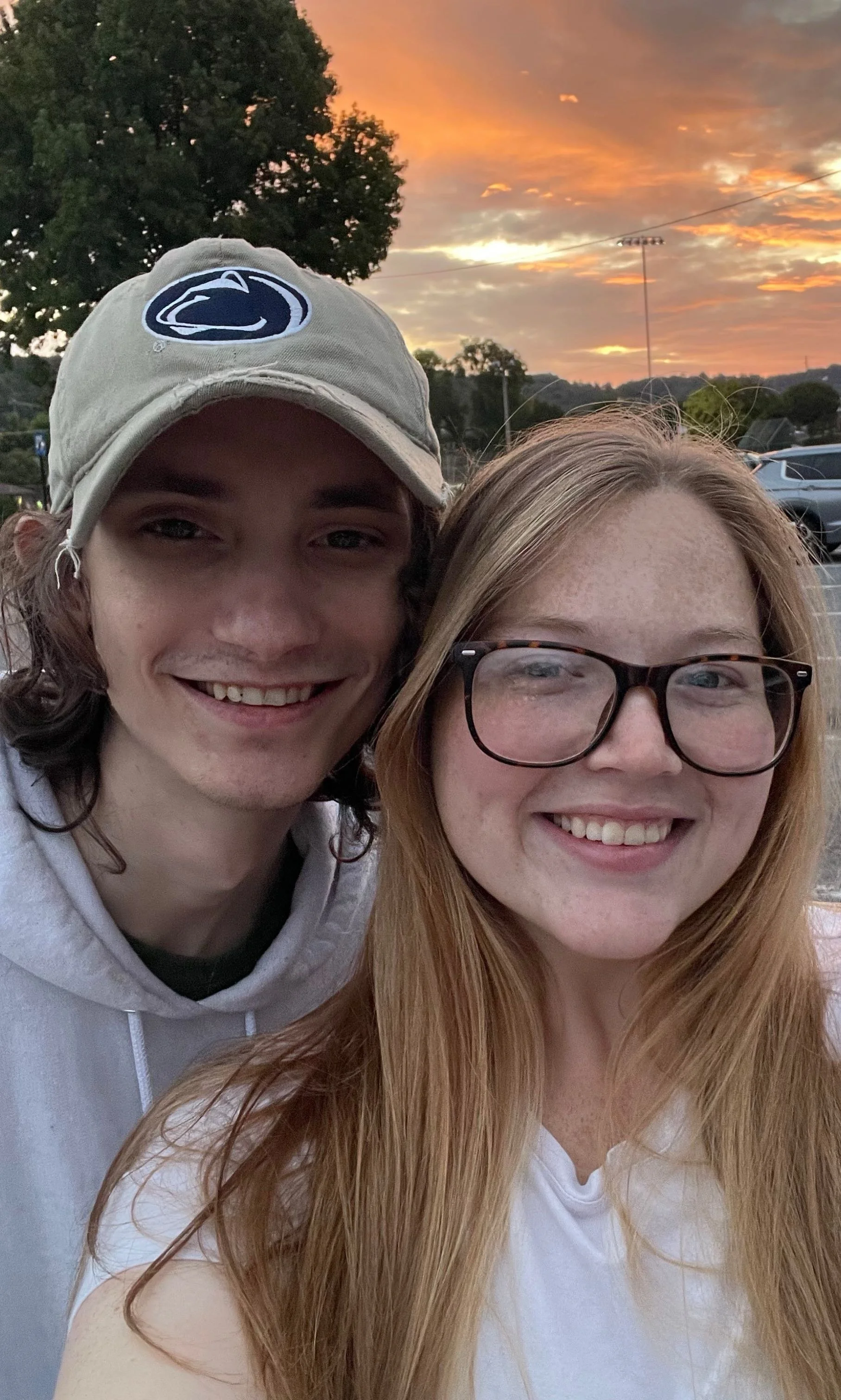 A young man and woman taking a selfie outdoors during sunset, with trees and parked cars in the background.