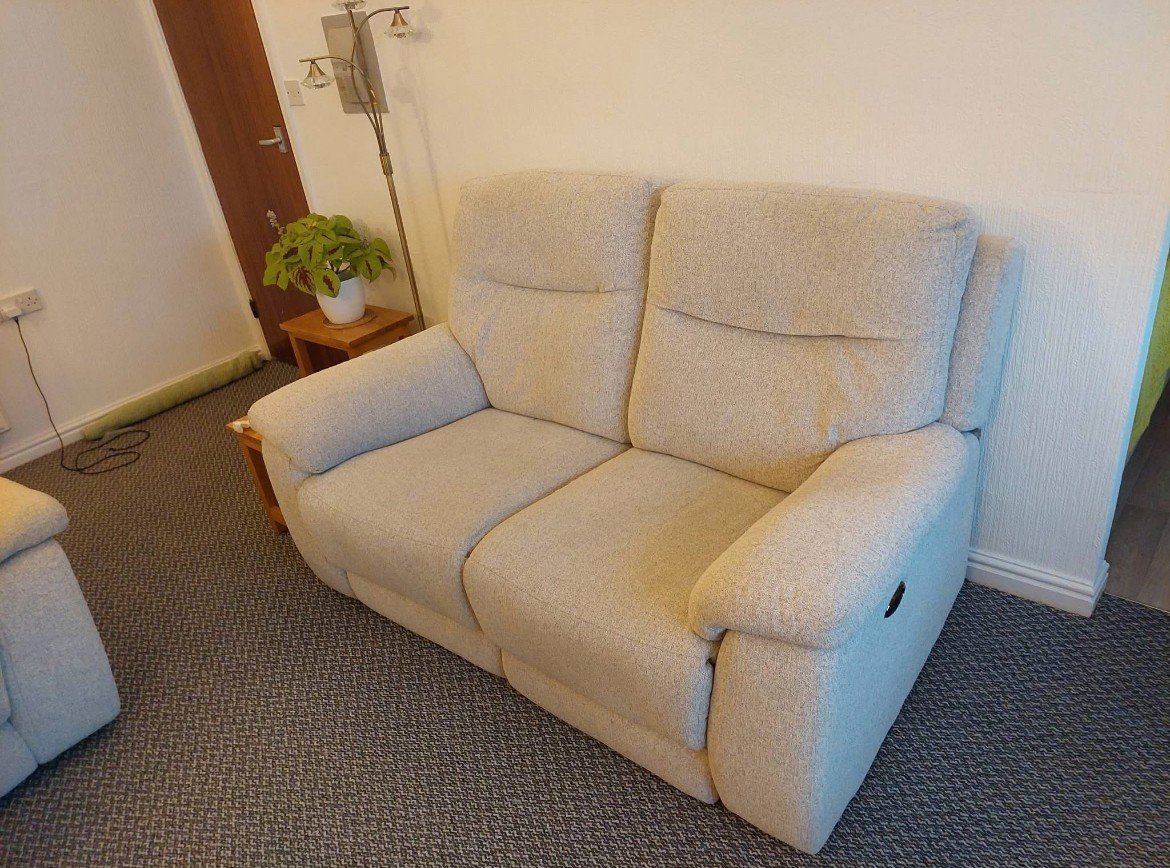Cream-colored fabric sofa in a living room with a small wooden side table and potted plant, beige walls, and dark patterned carpet.
