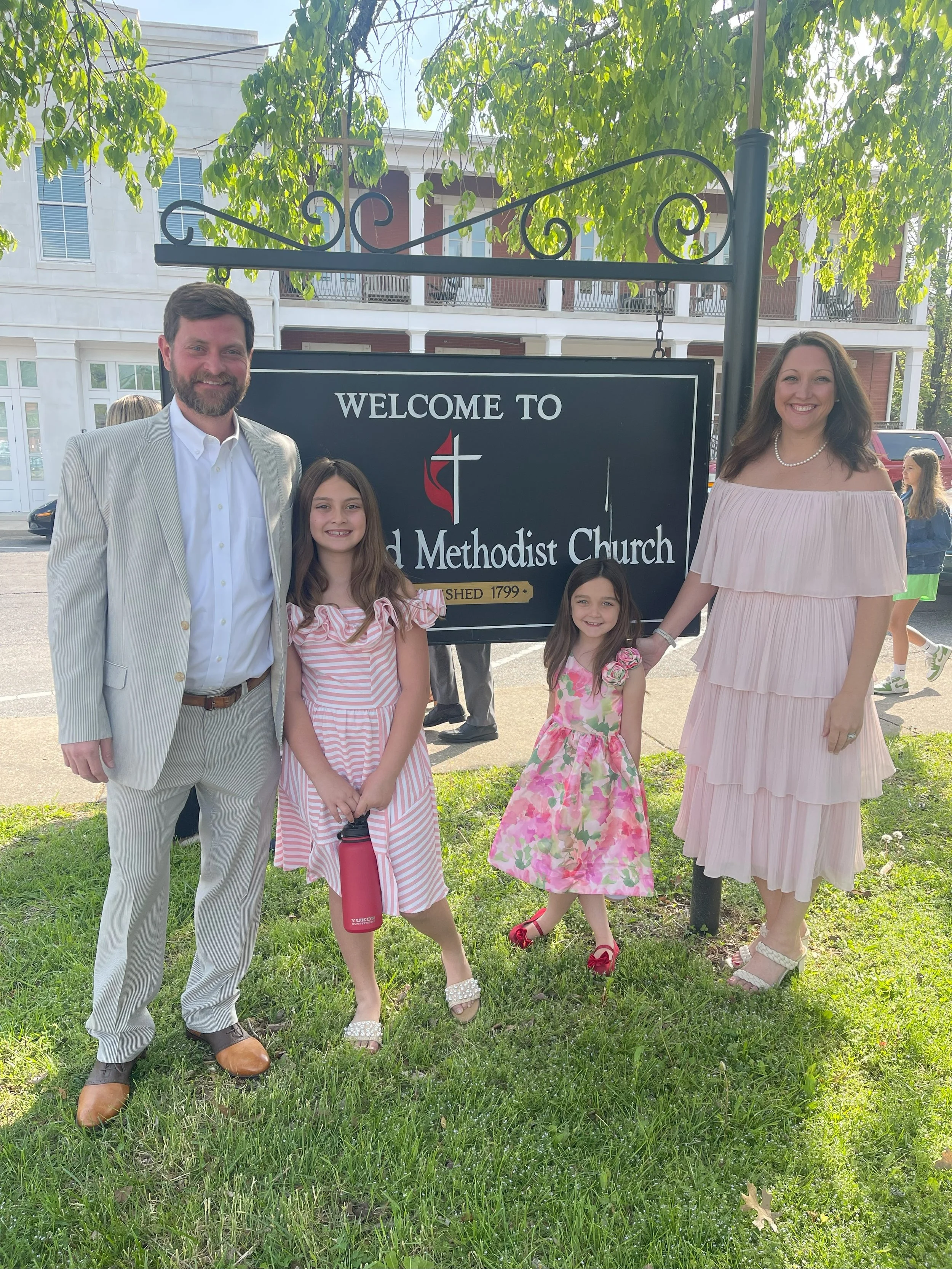 A family of four stands in front of a church sign, dressed in semi-formal attire, posing for a photo on a sunny day.