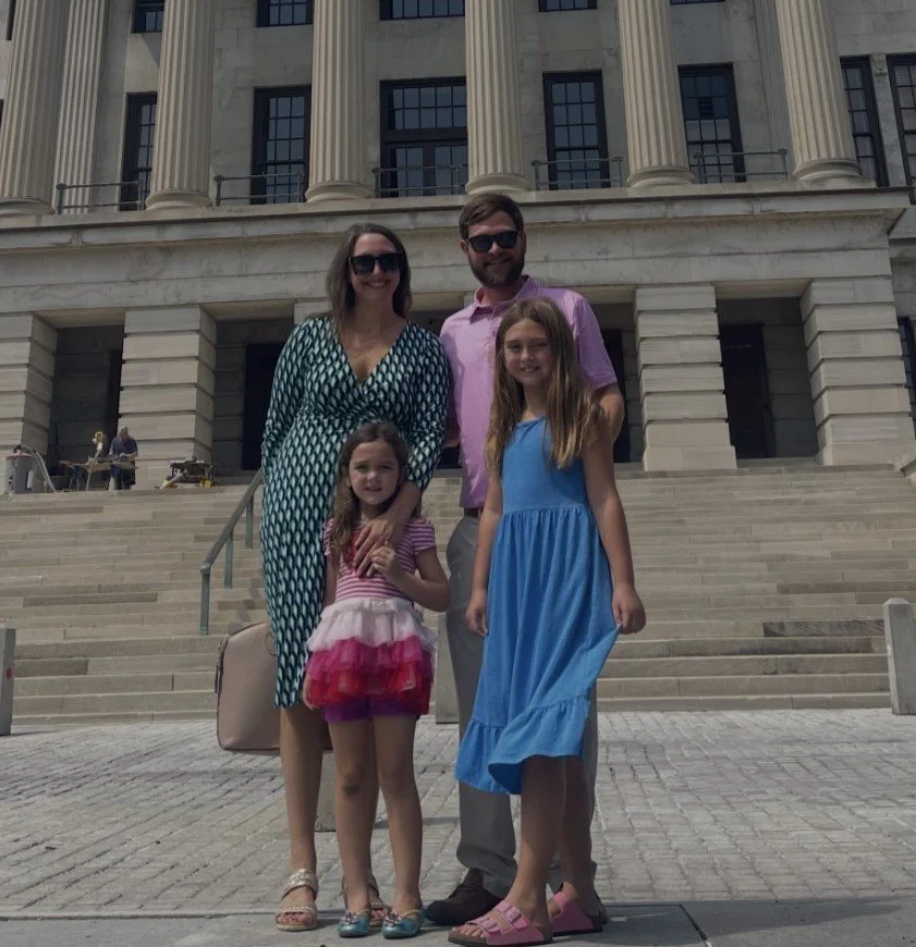 A family of four standing on stairs outside a large, classical stone building with tall columns.