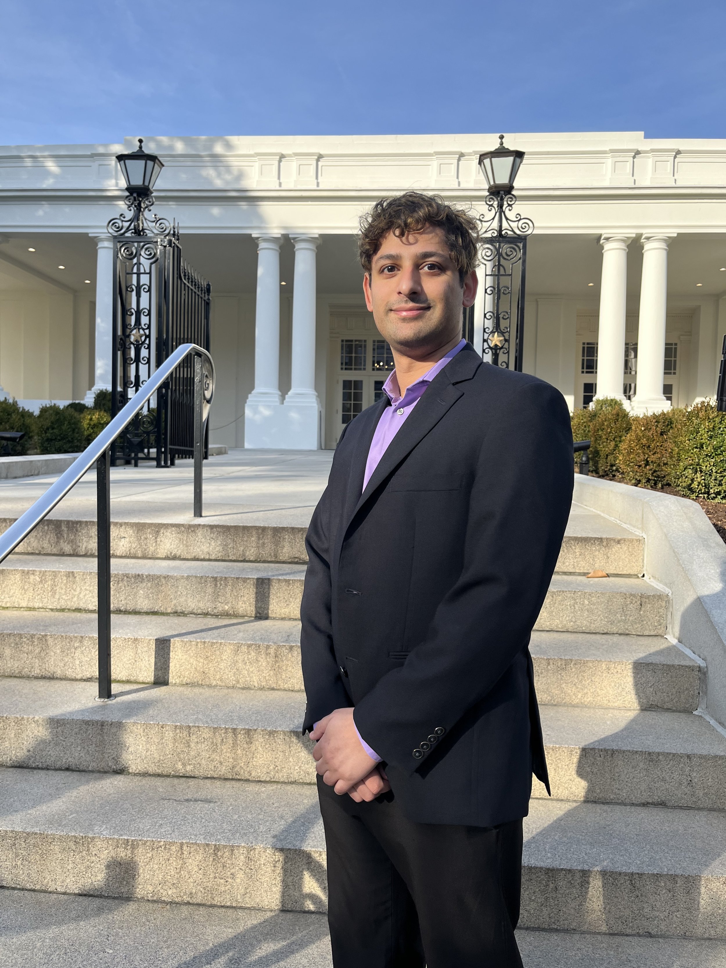 A young man in a black suit and purple shirt standing on front steps of a white building with columns and black wrought-iron gates.