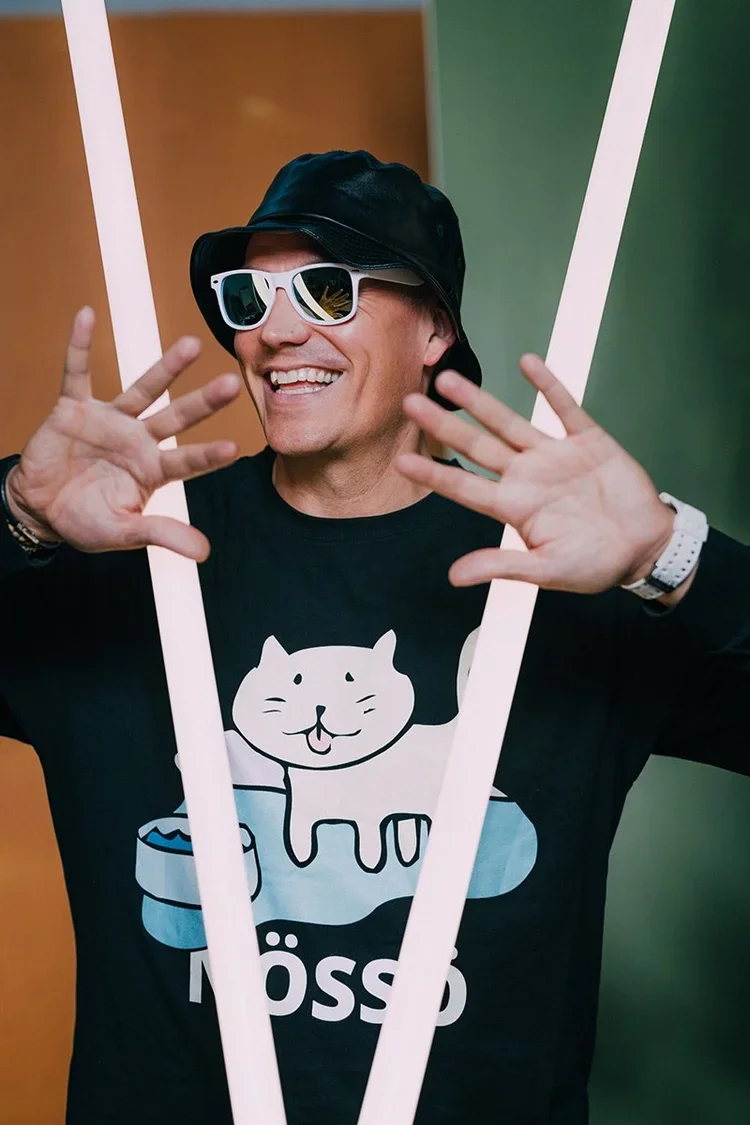 A smiling man wearing sunglasses, a black bucket hat, and a black T-shirt with a cat illustration and the word 'Möppo.' he is making a playful gesture with his hands, and is framed by two vertical white lines.