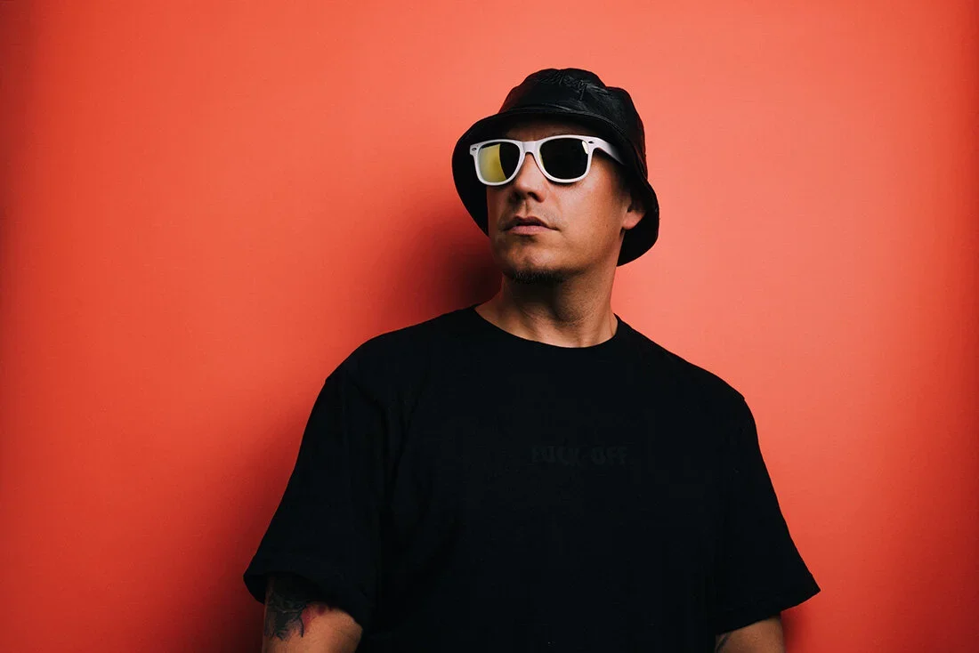 A man wearing white sunglasses, a black bucket hat, and a black t-shirt, standing against a pink wall.