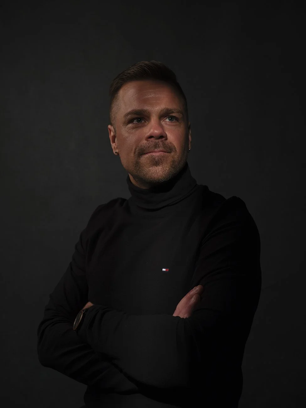 Portrait of a man with short brown hair and a beard, wearing a black turtleneck and standing against a dark background.