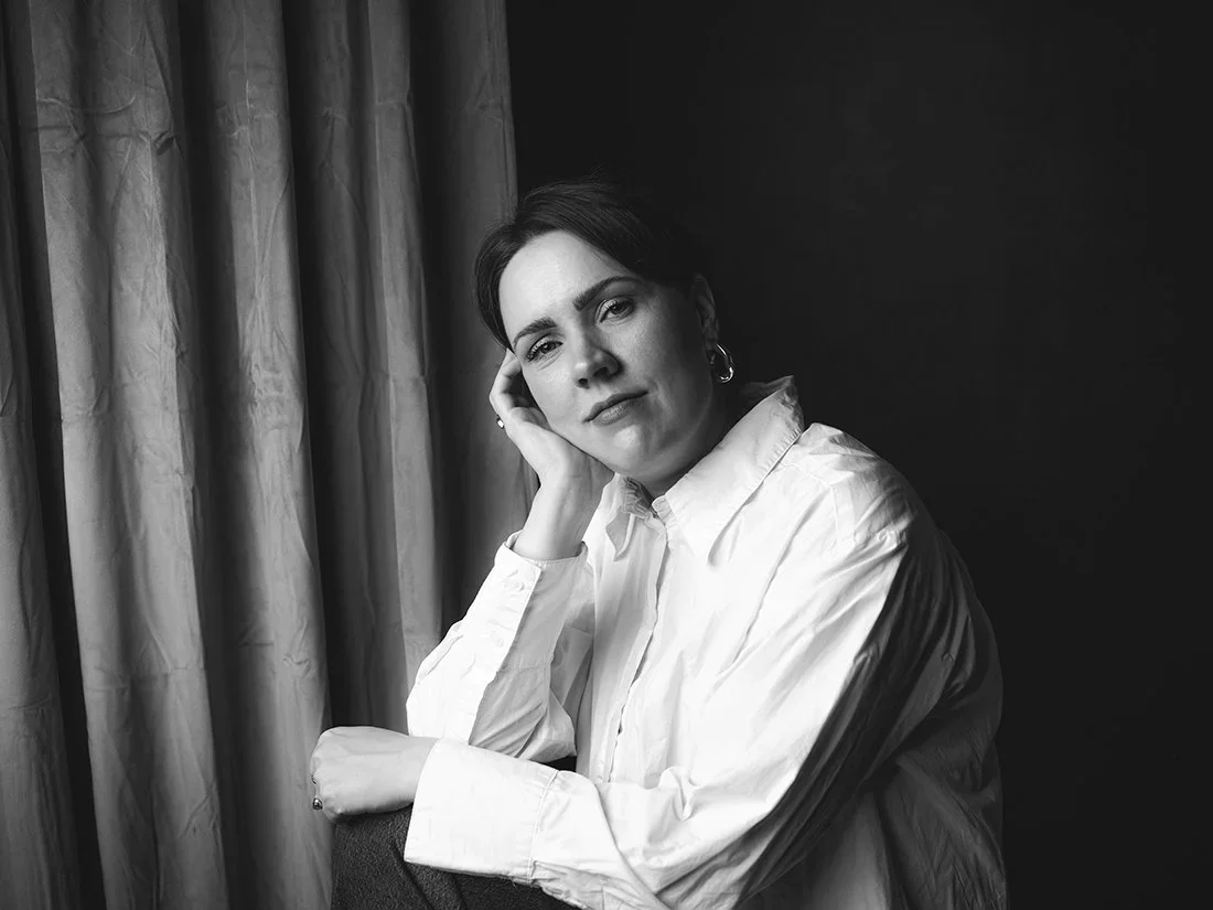 A woman posing for a black and white portrait, sitting with her head resting on her hand, wearing a white button-up shirt, and looking at the camera with a thoughtful expression.