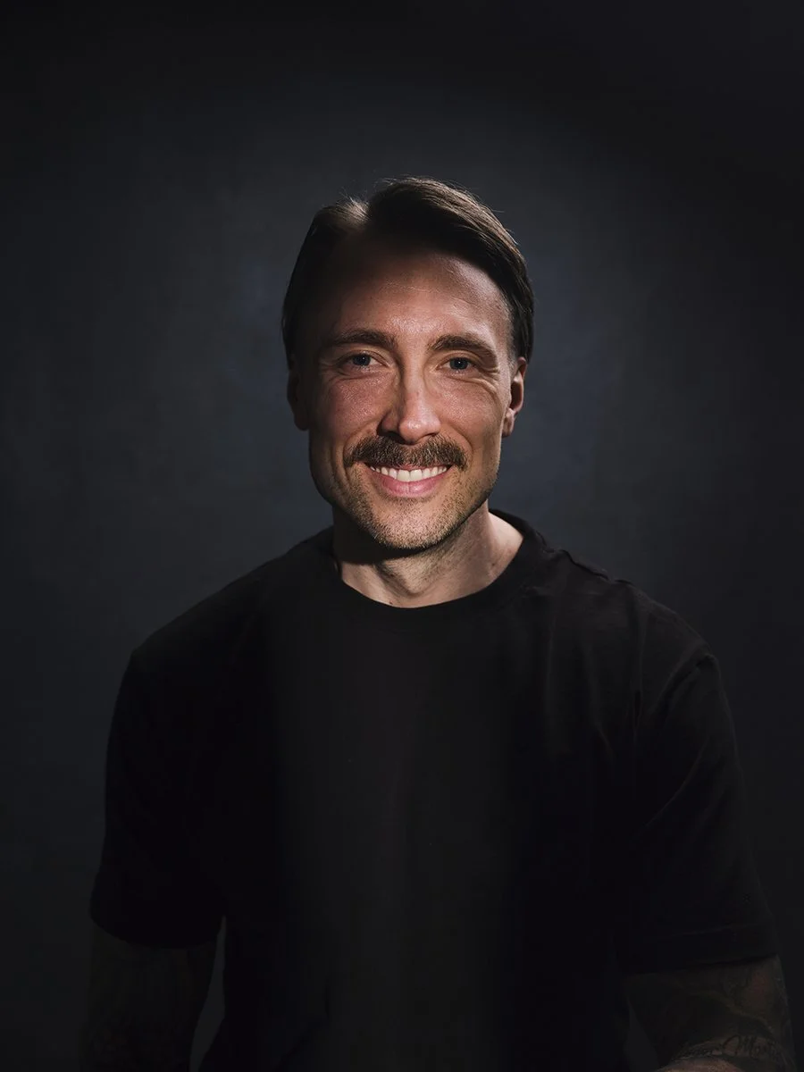 A smiling man with dark hair and a mustache, wearing a black shirt, against a dark background.