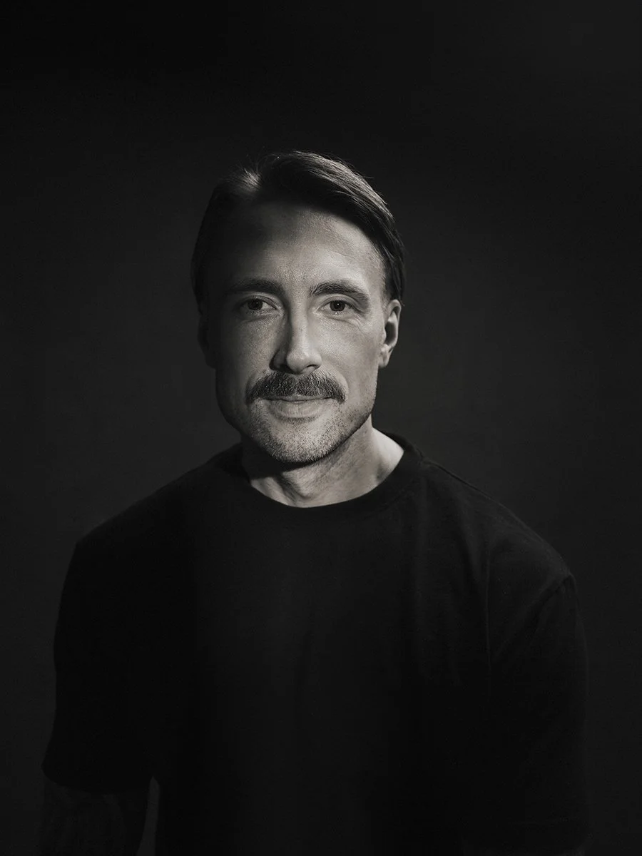 Black and white portrait of a man with a mustache and short hair, wearing a black shirt, against a dark background.