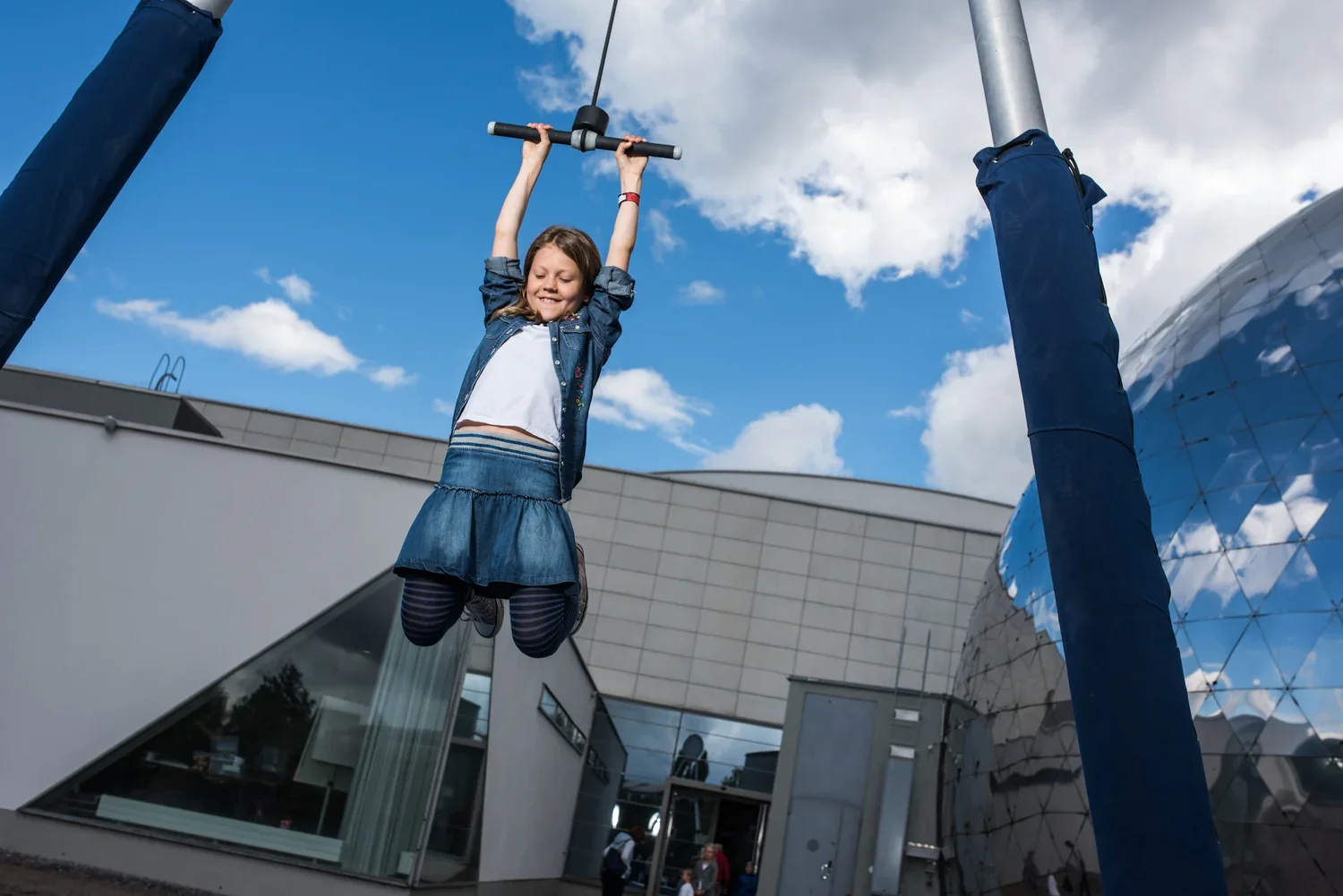 Girl in a denim jacket and skirt is hanging from a pull-up bar outdoors, with a modern building and partly cloudy blue sky in the background.