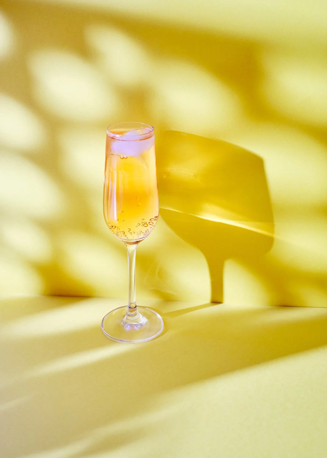 A champagne flute filled with bubbly, pink-tinted champagne, casting a pink shadow on a yellow background.