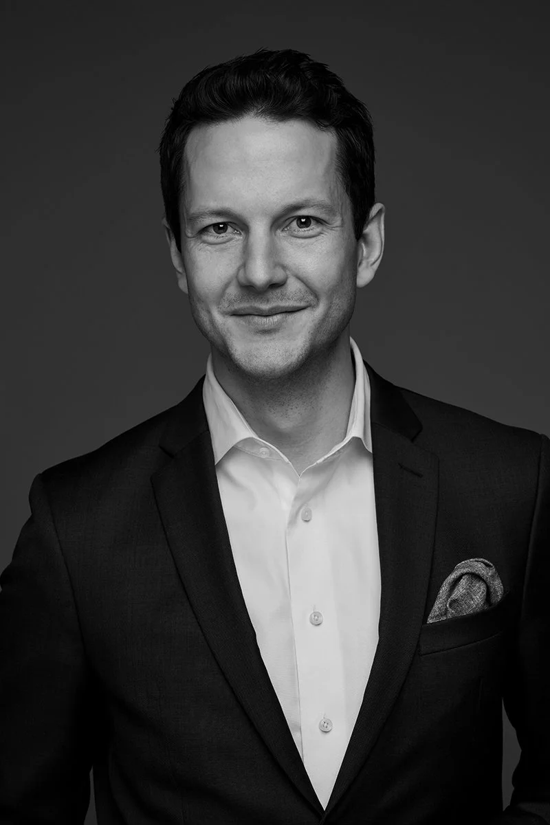 A black and white portrait of a man in a suit with a white shirt and pocket square, smiling at the camera.