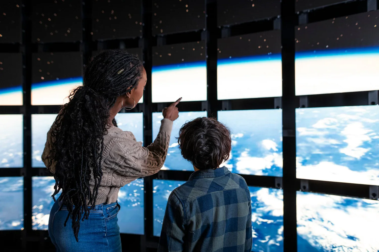 A woman and a boy in front of multiple screens displaying images of Earth from space, with the woman pointing at one of the screens.