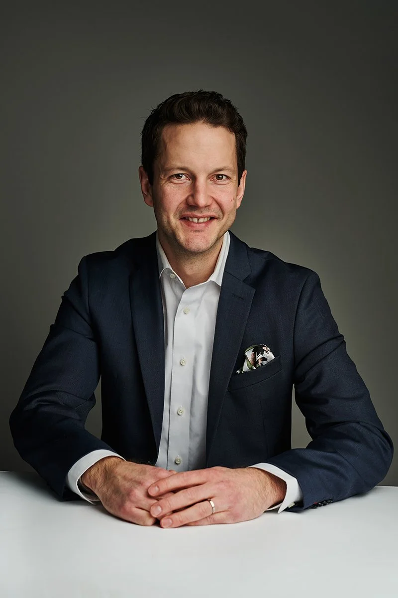 Portrait of a man in a dark suit and white shirt, smiling, sitting at a white table against a plain gray background.