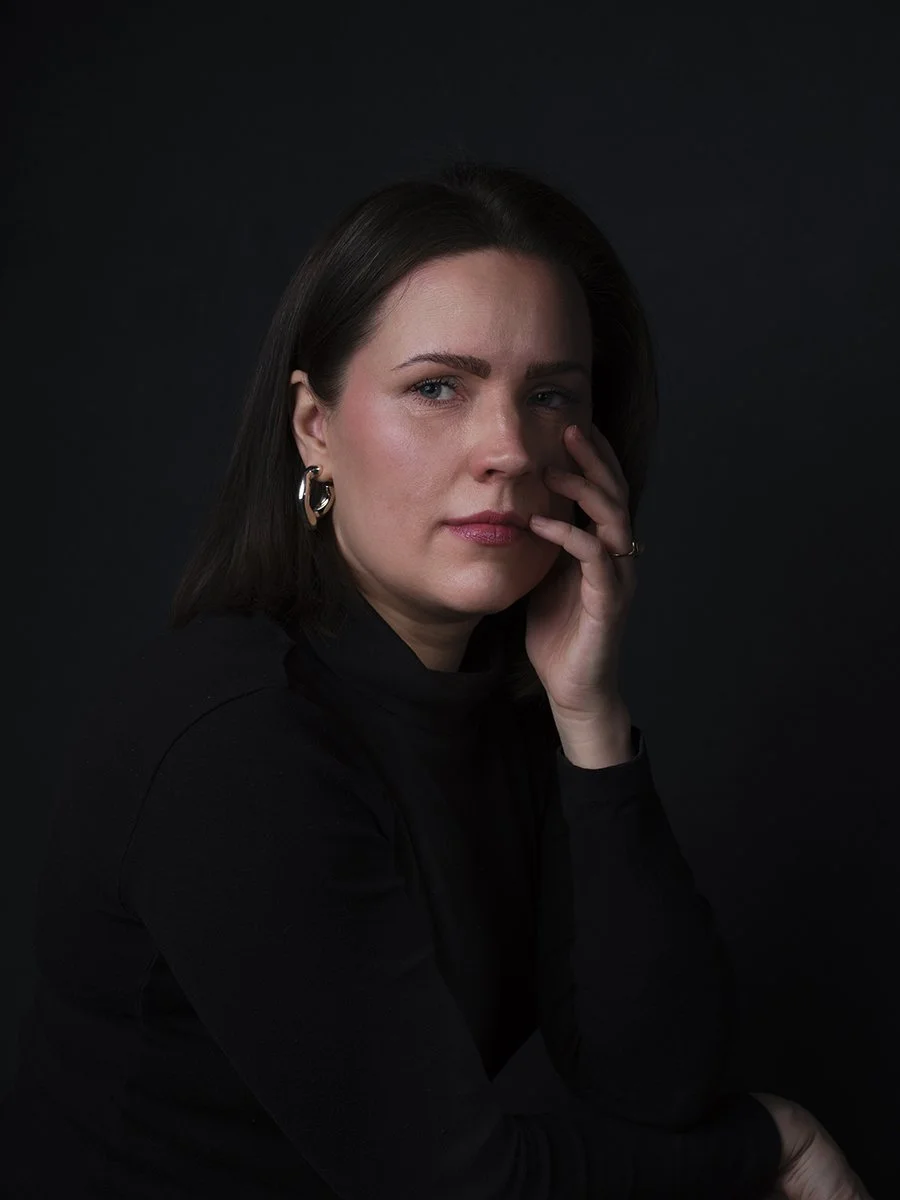 A portrait of a woman with dark hair wearing a black turtleneck and hoop earrings against a black background.