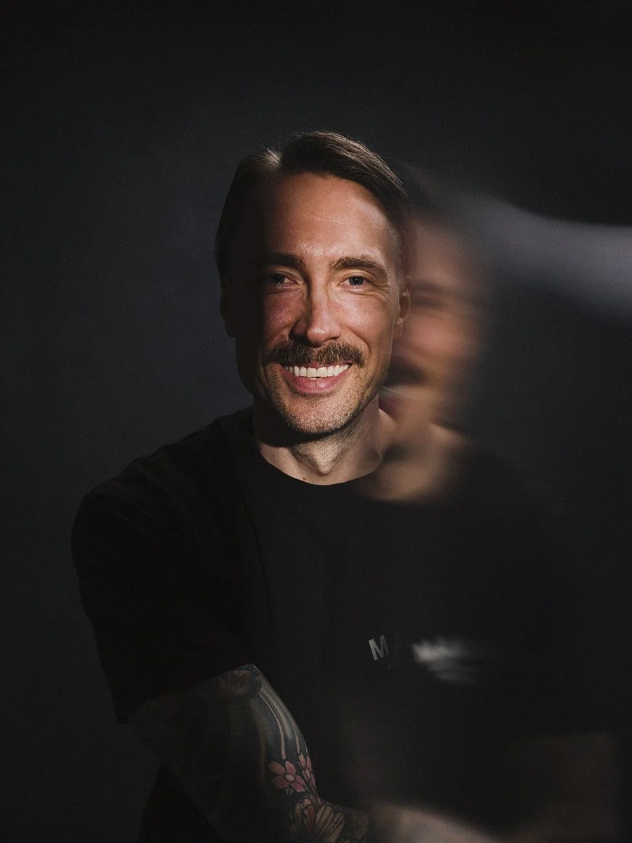 A smiling man with short brown hair, a beard, and tattoos on his arm, wearing a black shirt, against a dark background with a blurred second image of himself in motion.