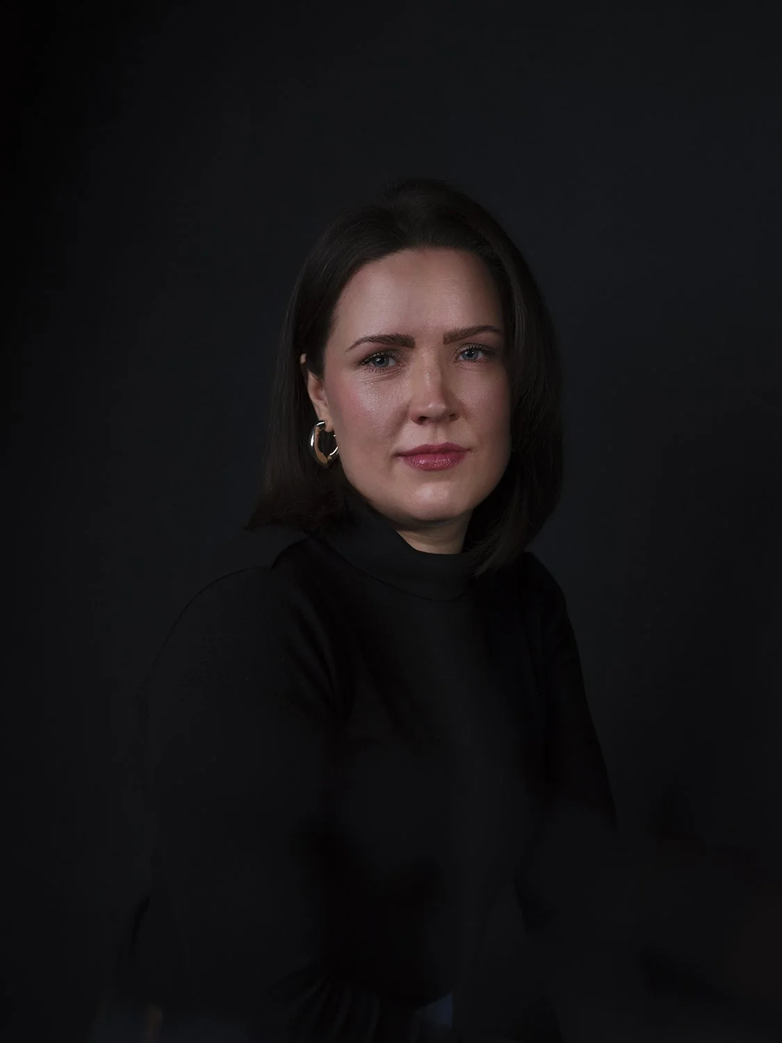 A woman with dark hair and blue eyes poses against a black background, wearing a black turtleneck and gold hoop earrings.
