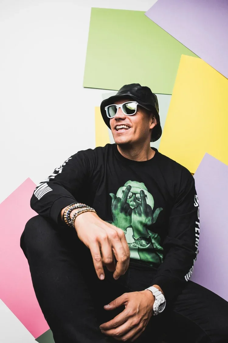 A smiling man wearing sunglasses, a black bucket hat, a graphic black sweatshirt, and multiple bracelets, sitting in front of a colorful background of pastel rectangles.