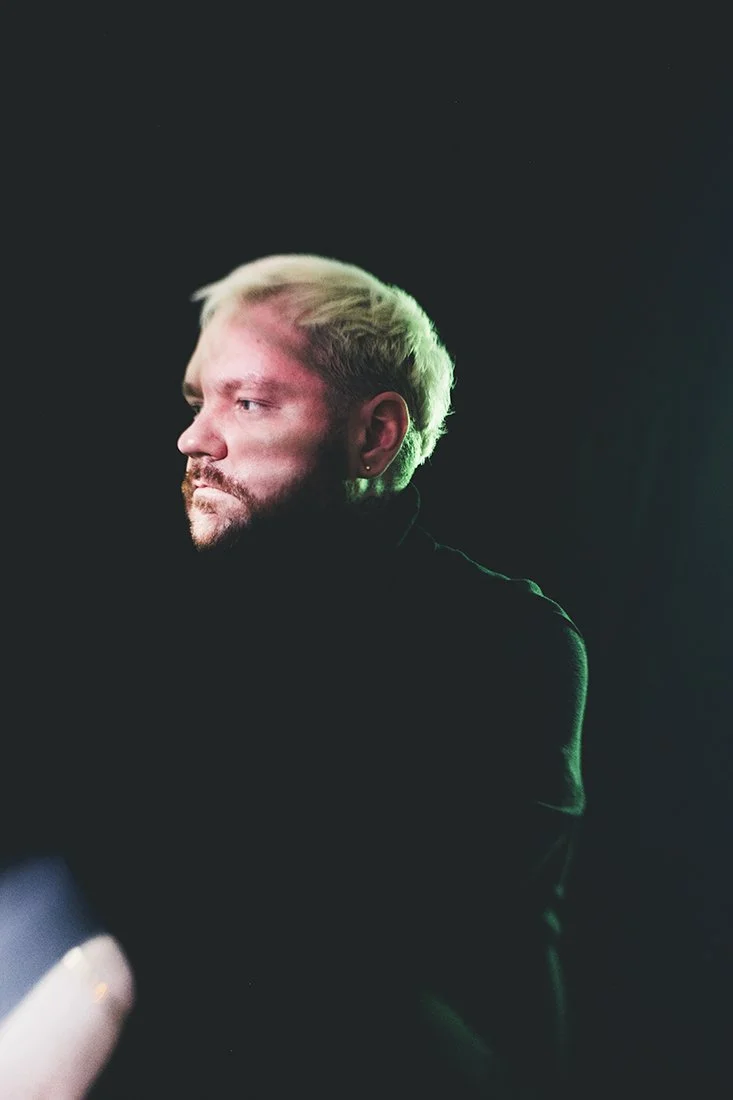 Profile of a man with blond hair and a beard, wearing a black shirt or jacket, against a dark background.