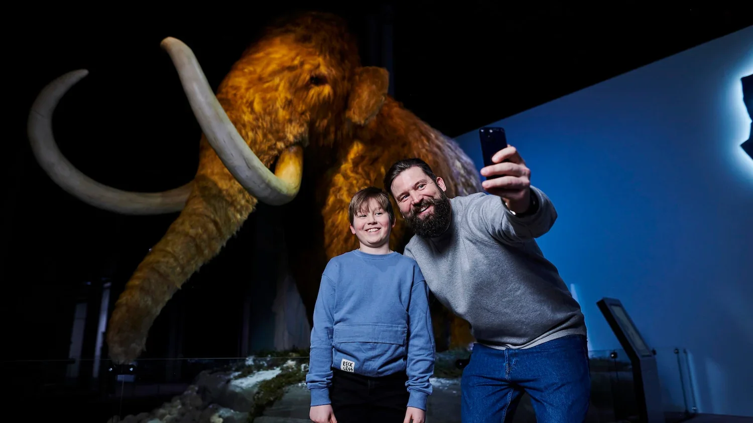 A man and a boy taking a selfie in front of a large woolly mammoth exhibit at a museum.