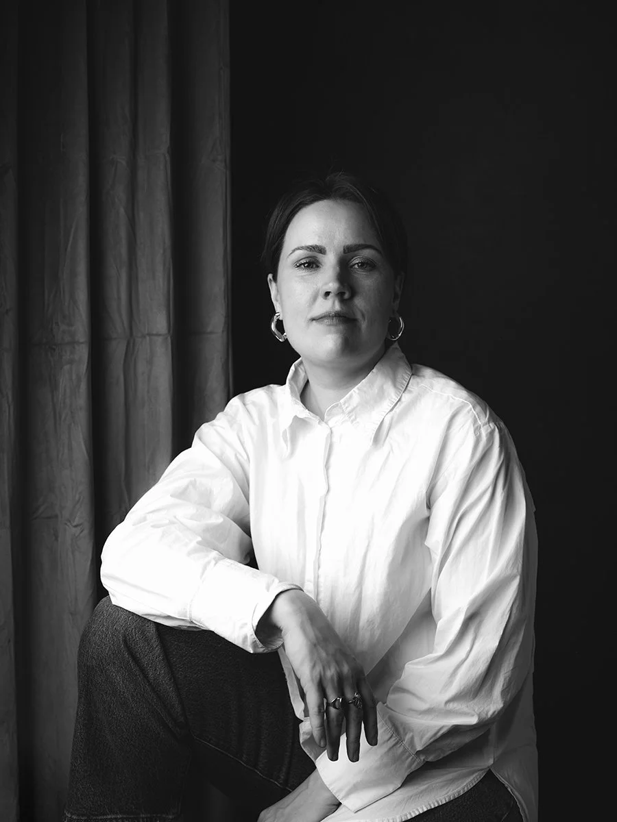 Black and white portrait of a woman with short dark hair, wearing a white button-up shirt and hoop earrings, sitting with her hand resting on her knee in front of a dark background and a curtain.