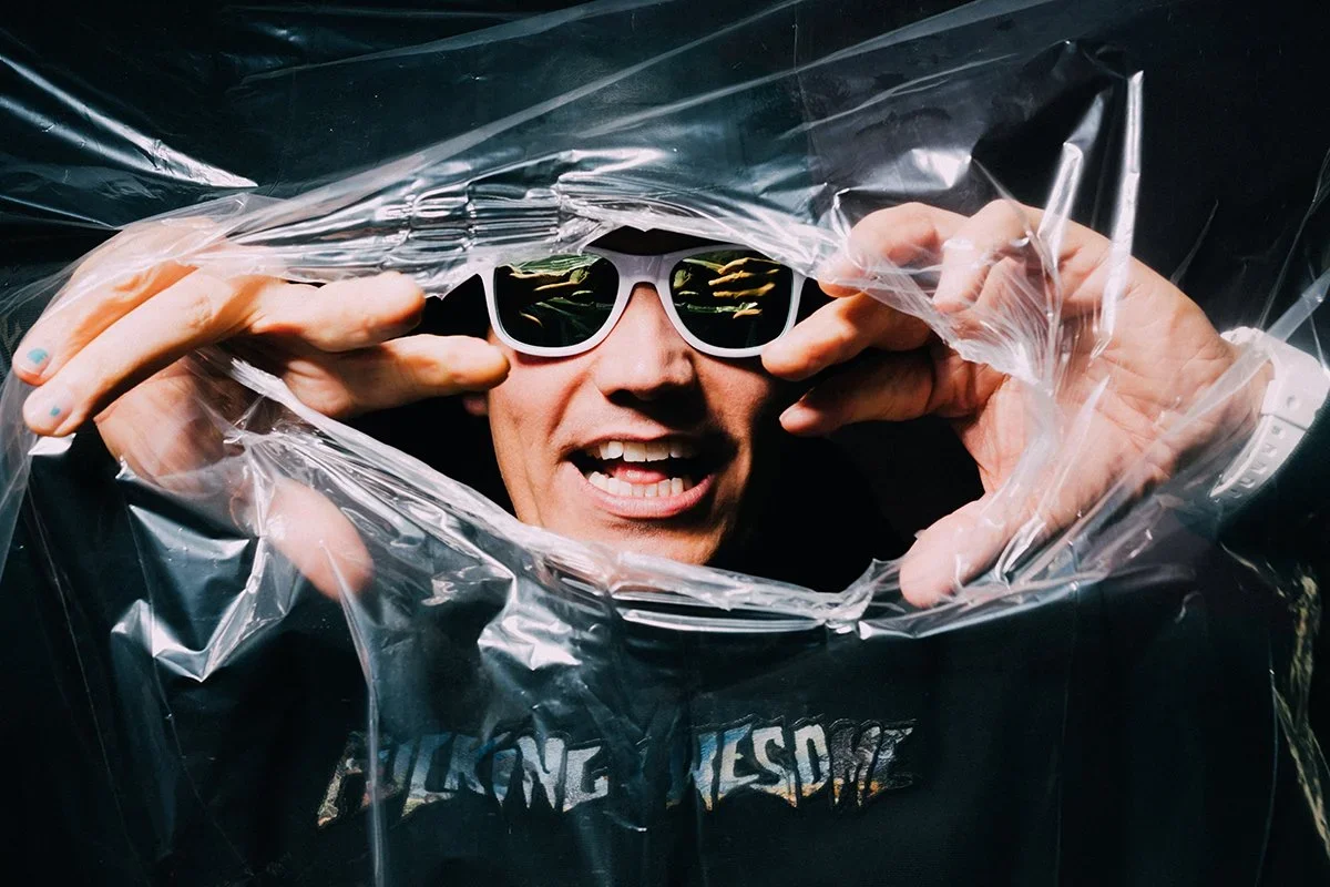 Man wearing sunglasses and a black hoodie, peering through clear plastic, making a playful expression.