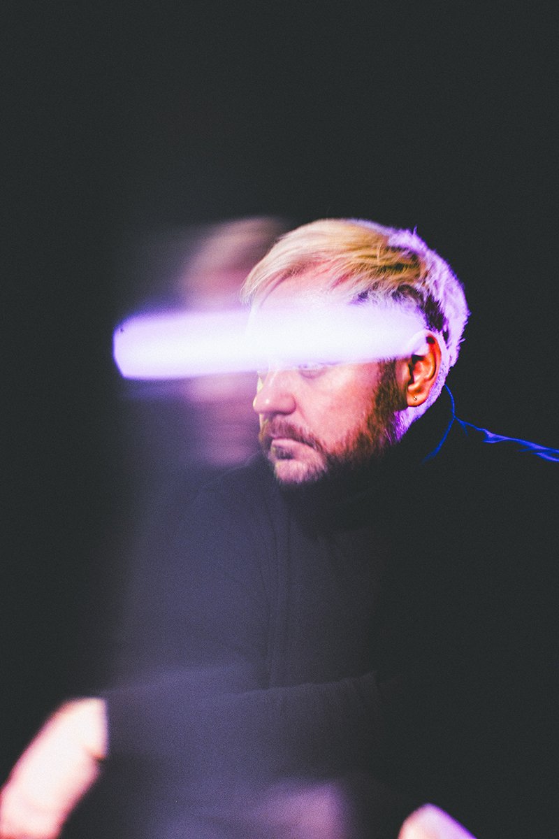 A person with blond hair and a beard looking to the side, illuminated by horizontal purple and white light streak across their eyes.