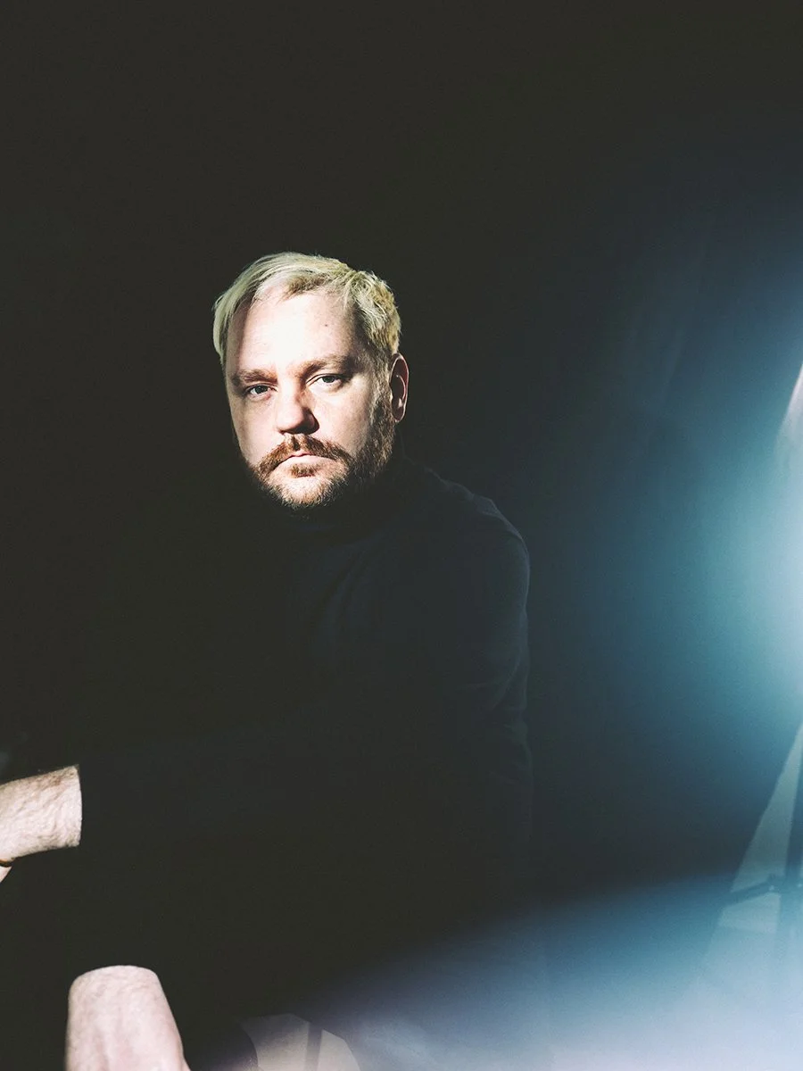 A man with blond hair and a beard, wearing a black shirt, sitting in a dark room, illuminated by a soft light source on the right.