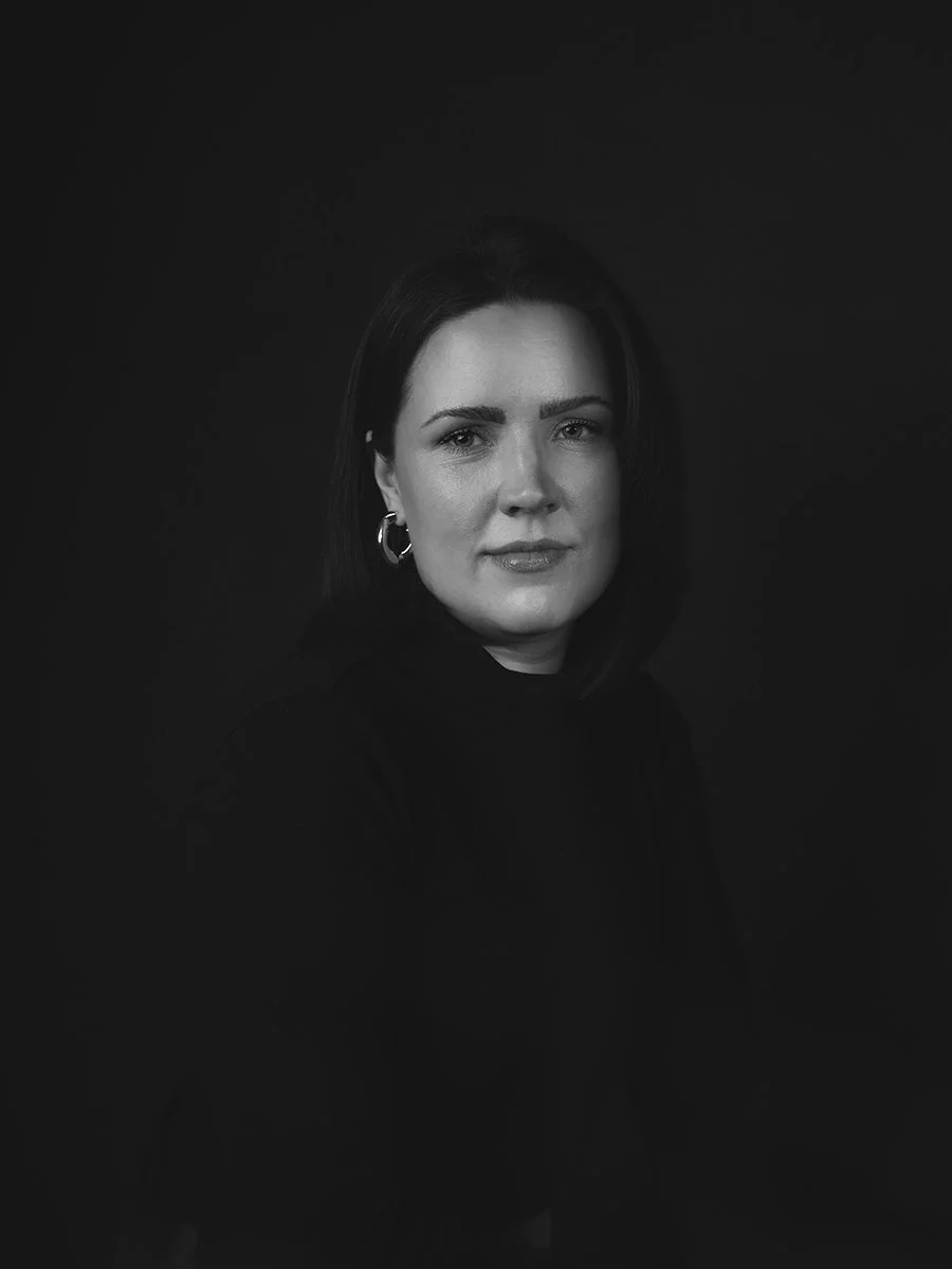 Black and white portrait of a woman with shoulder-length dark hair, wearing hoop earrings, and looking directly at the camera against a black background.