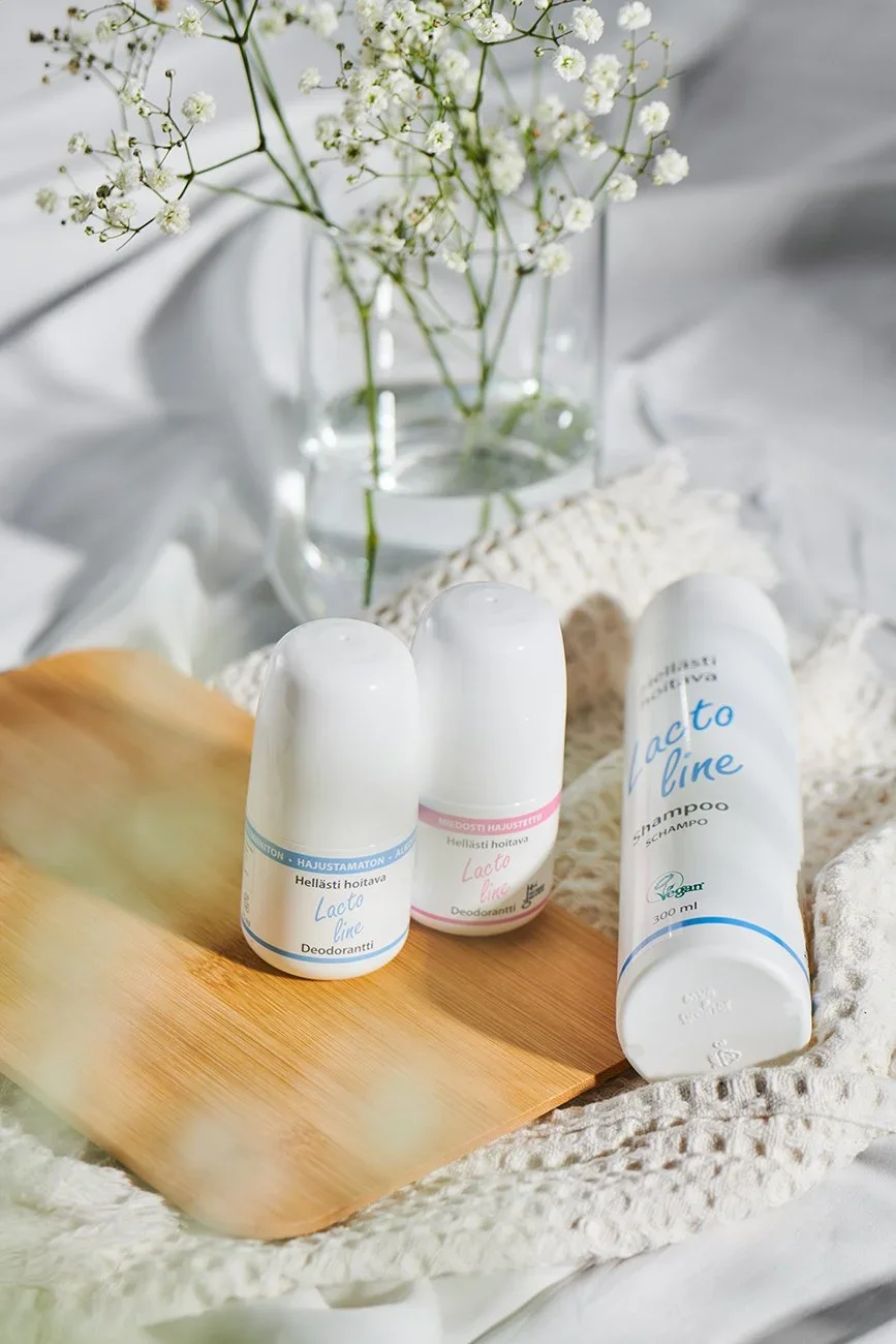 A display of three Lactoline deodorant bottles and a bottle of Lactoline shampoo on a wooden board, with a white lace cloth underneath. In the background, there is a glass vase with white baby's breath flowers, all arranged on a white fabric surface.