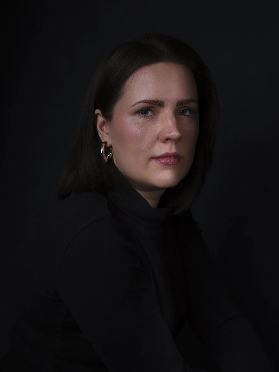 A portrait of a woman with dark hair, blue eyes, pierced ear jewelry, wearing a black turtleneck, against a black background.