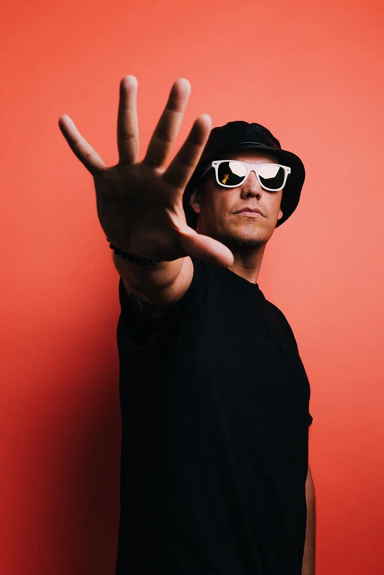 Man wearing sunglasses and a bucket hat, reaching out with his hand towards the camera, standing against a pink background.