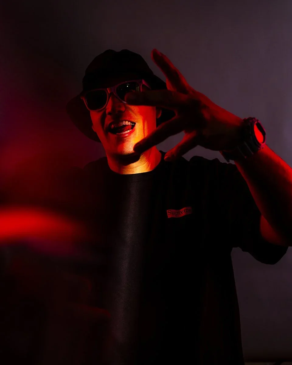 Person wearing sunglasses and a black hat, smiling and making a peace sign gesture with their hand, in low lighting with red accents.
