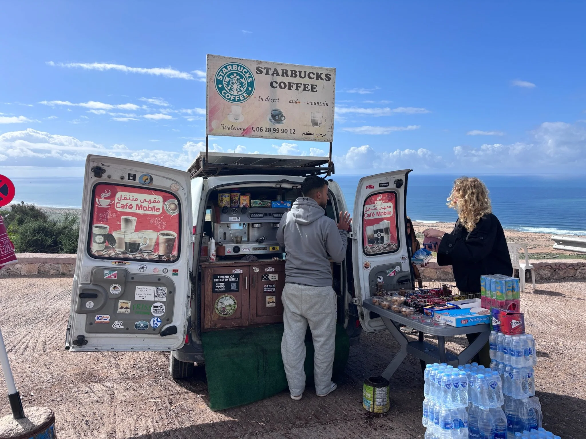 En mobil kaffevogn med mennesker, der handler kopper kaffe, med oceanen og en blå himmel i baggrunden.
