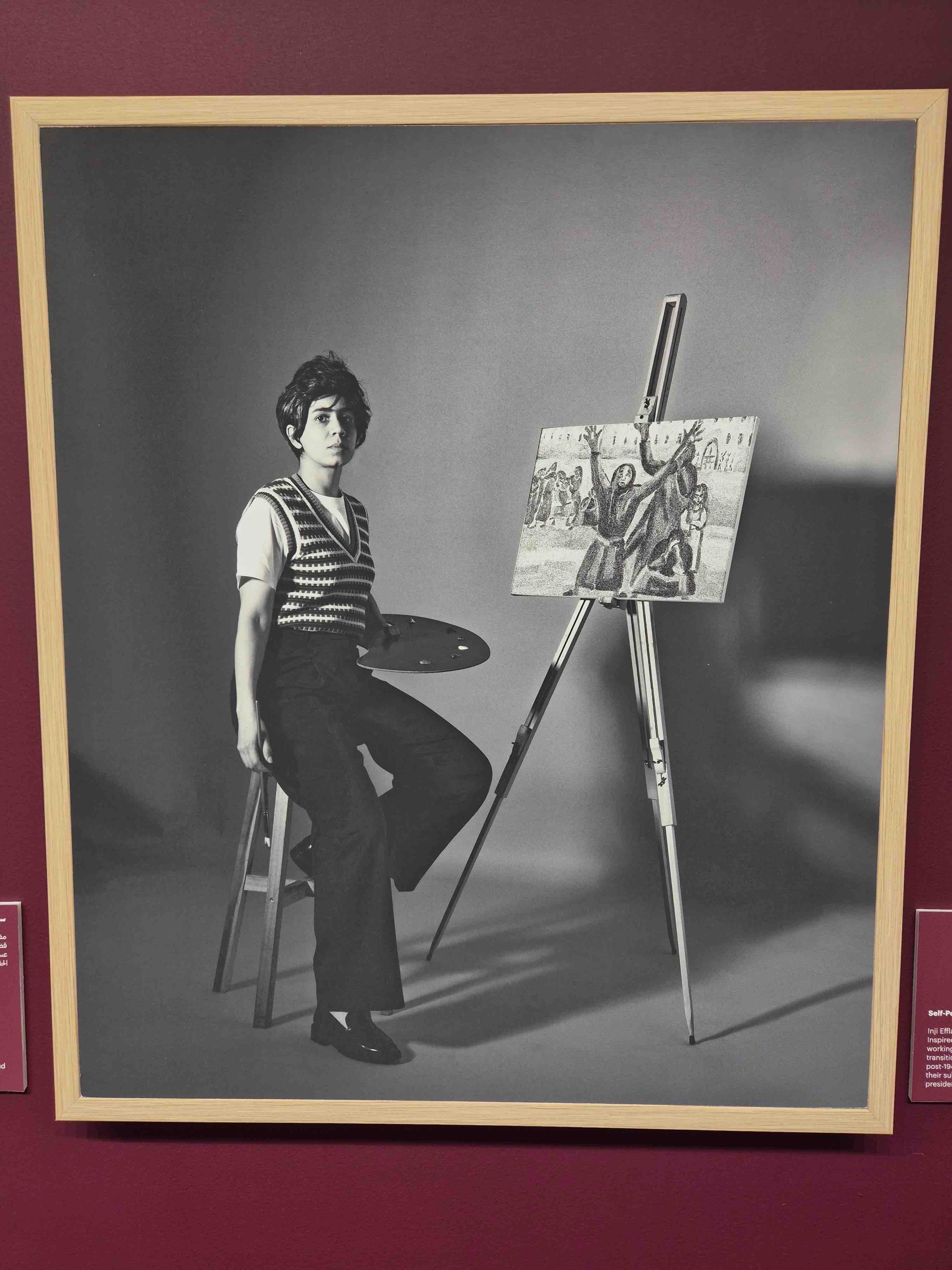 Black and white photograph of a woman sitting on a stool with an artist's palette, positioned next to an easel with a painting of a group of people, including some with raised arms, in an outdoor setting.