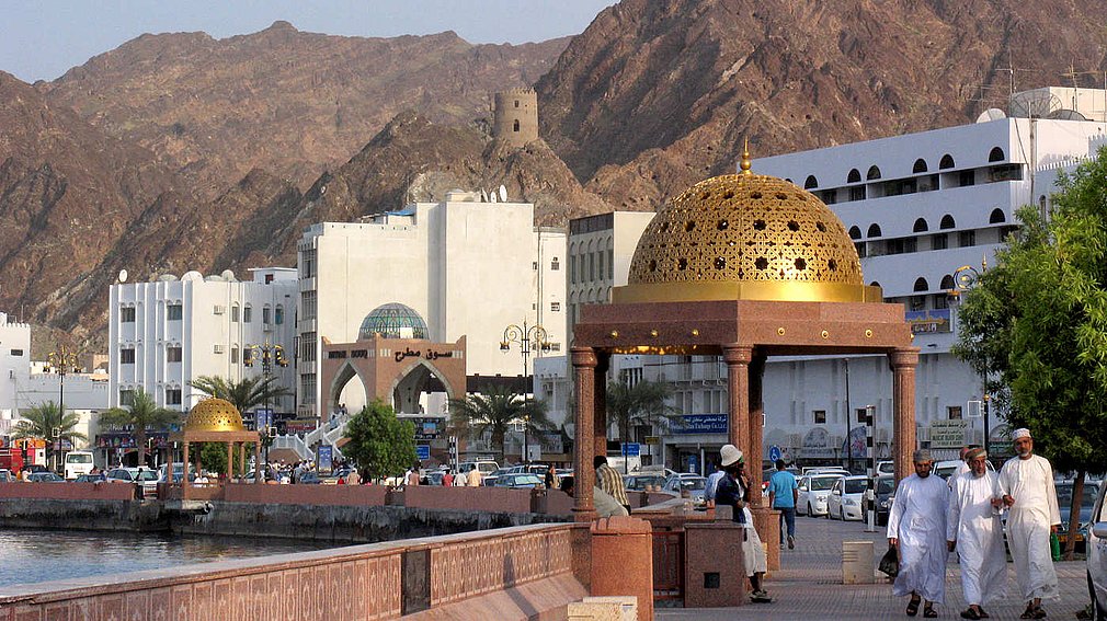 A waterfront scene in Muttrah Oman with white buildings, mountains, and a prominent golden dome structure, with people walking along the promenade.