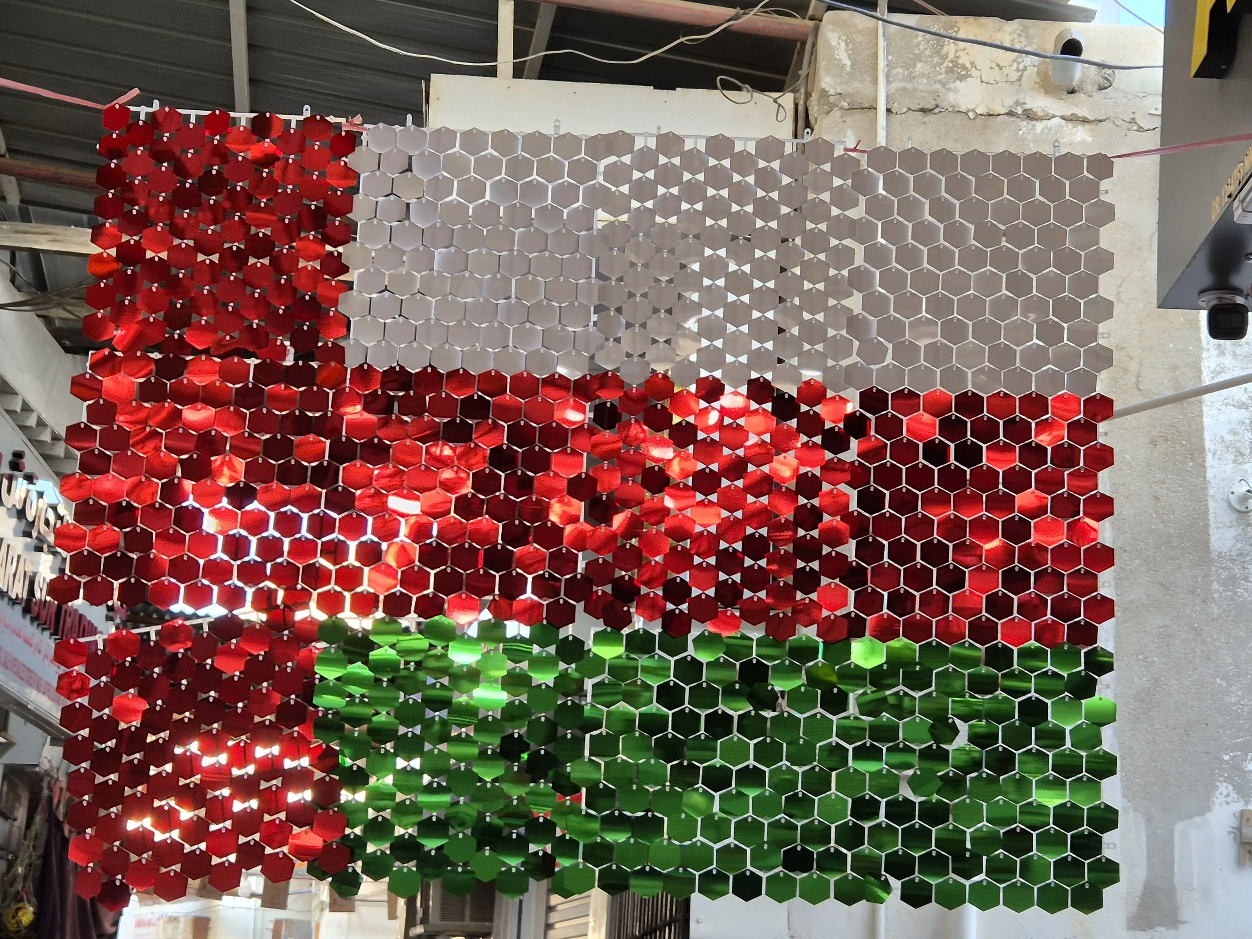 Colourful Omani mosaic art piece with red, green, gray hexagonal tiles, hung inside a building with a roof and some electrical wiring visible.