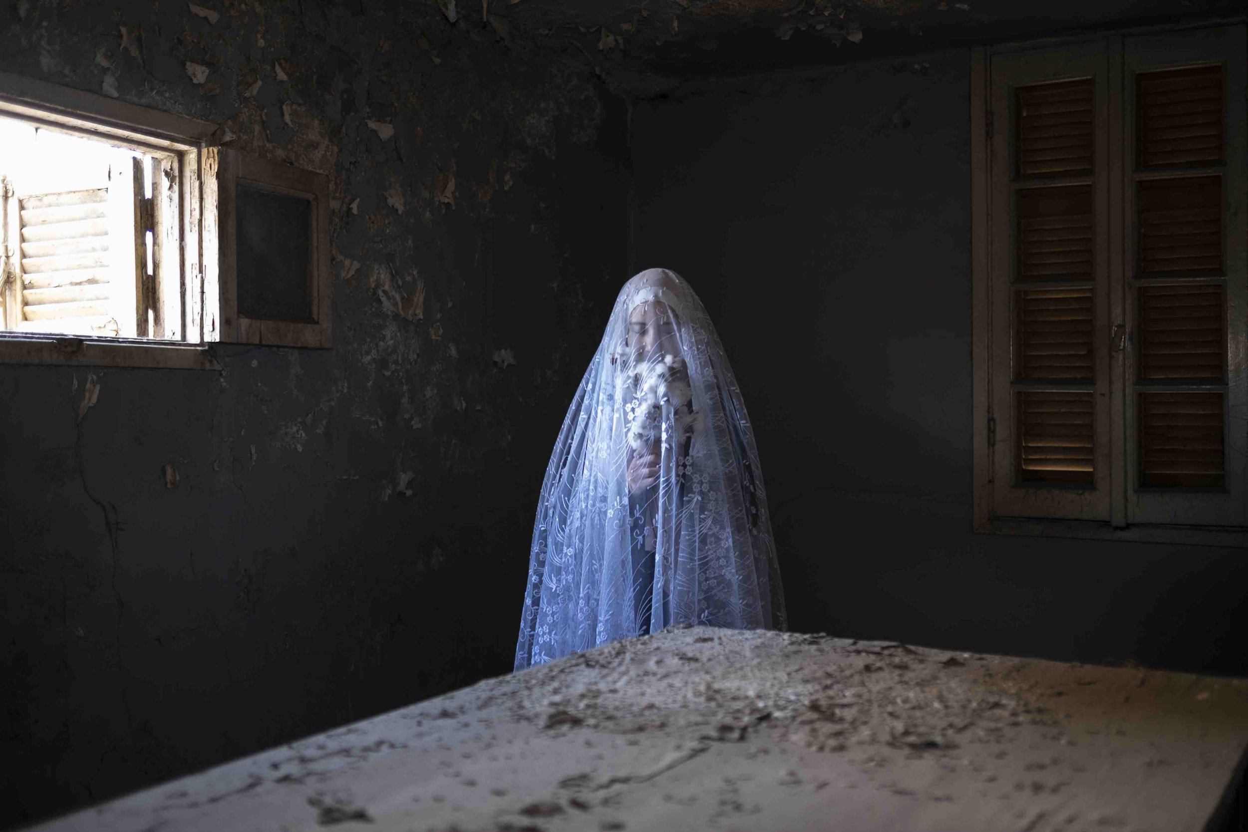 Amina Kadous' image of a  woman dressed in white holding a bouquet, wearing a veil, standing in a dark, abandoned room with peeling walls, a window with broken blinds, and dirty furniture.