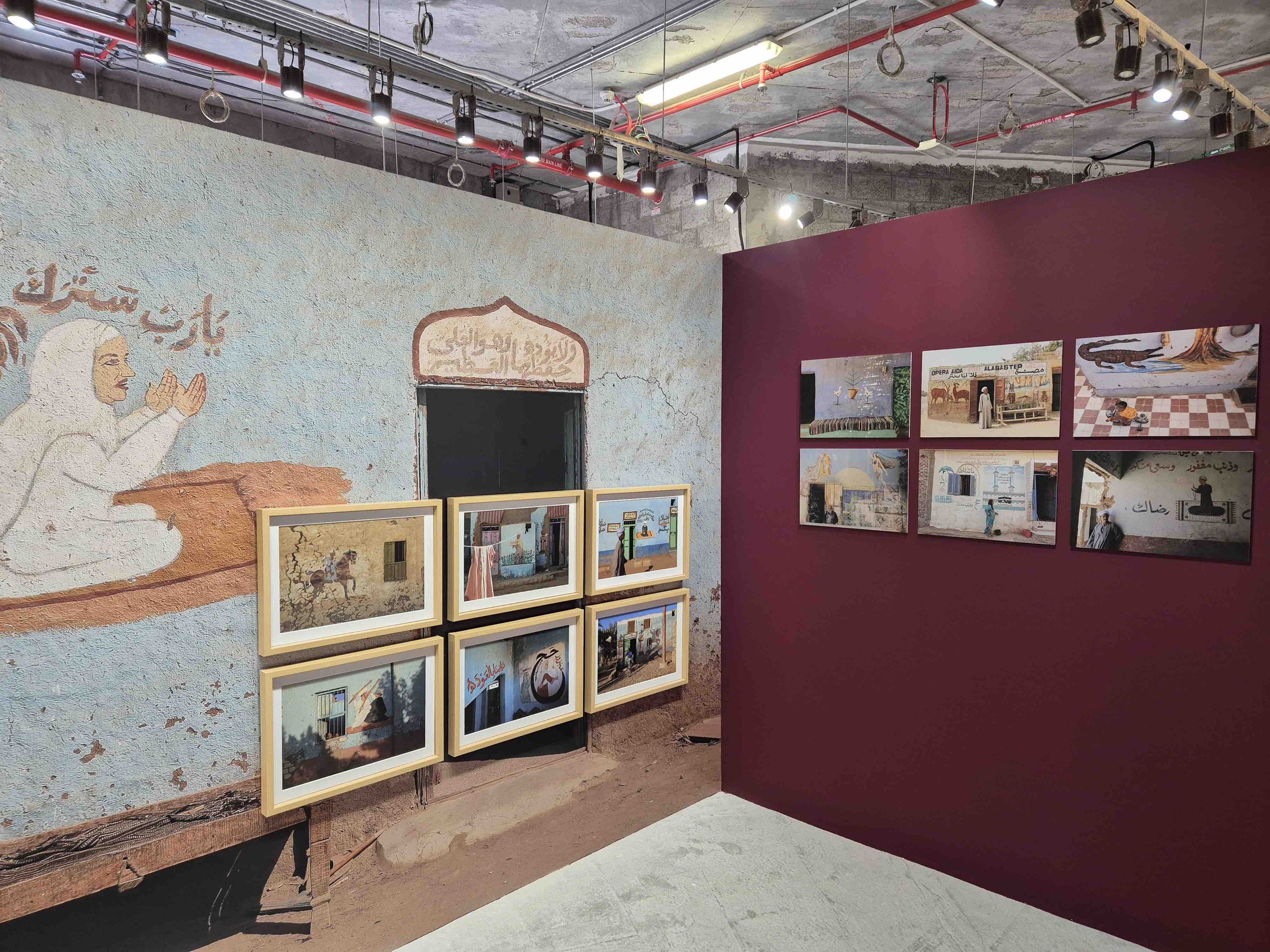 Egyptian artist Wafaa Samir's photographs displayed on a purple wall and a mural painted on an adjacent wall. The mural features Arabic writing and a woman with white hair in traditional clothing.