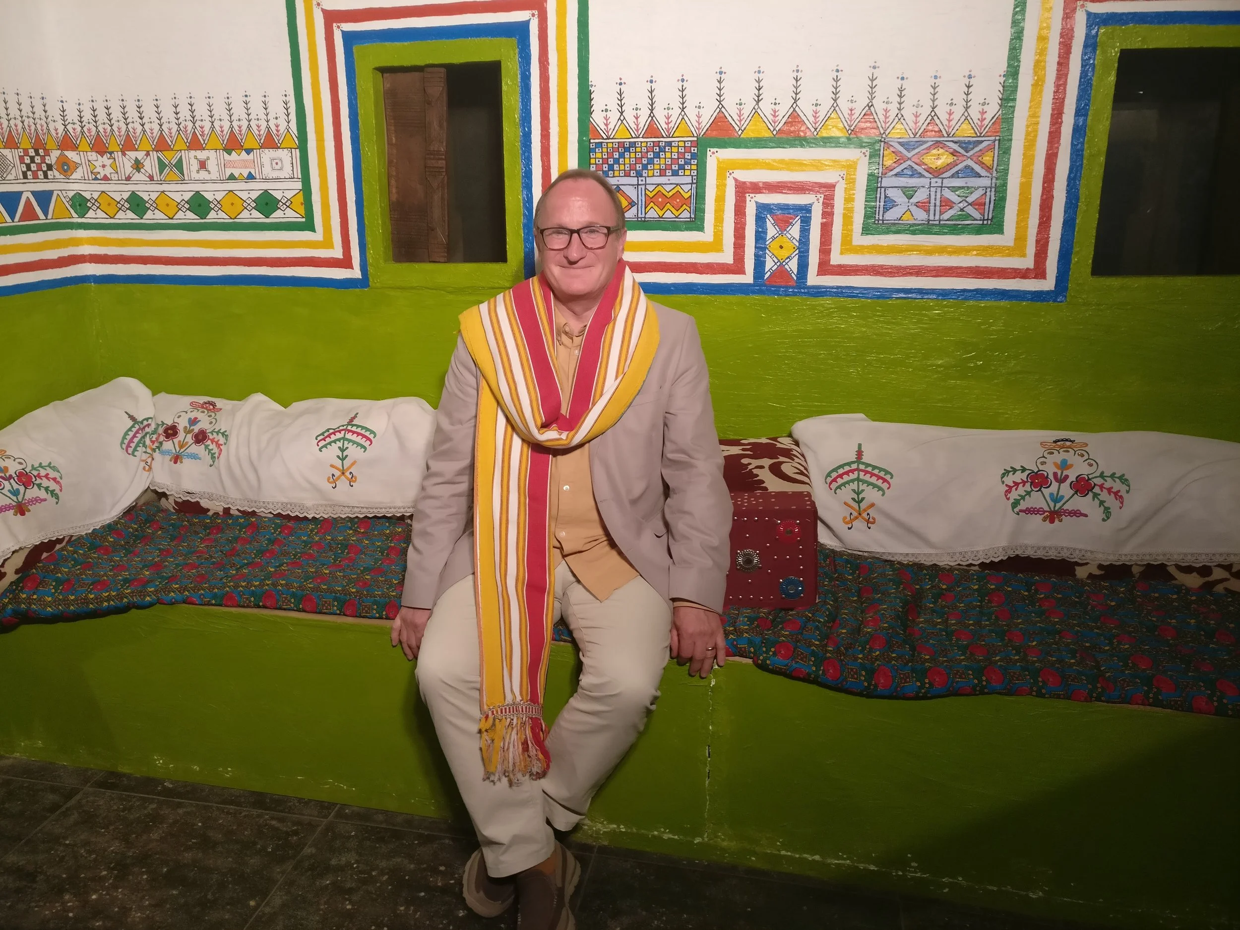 David Drake sitting on a colorful, traditional bench in a room with vibrant, decorated walls and embroidered cushions.