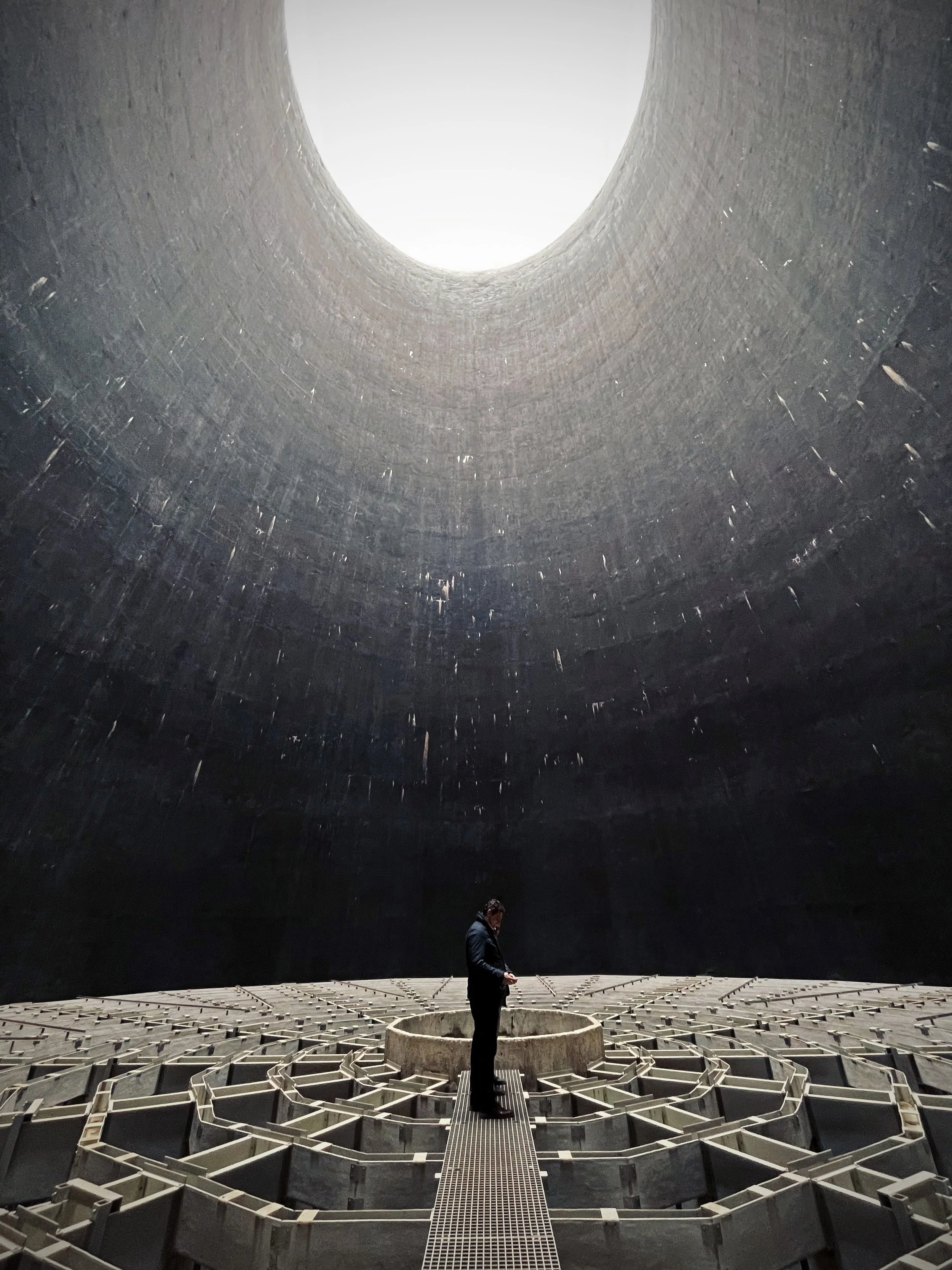 An image by artist Amina Kadous of a person standing inside a large, dark, industrial structure with a circular opening at the top allowing light in, and a patterned floor with a central circular element.
