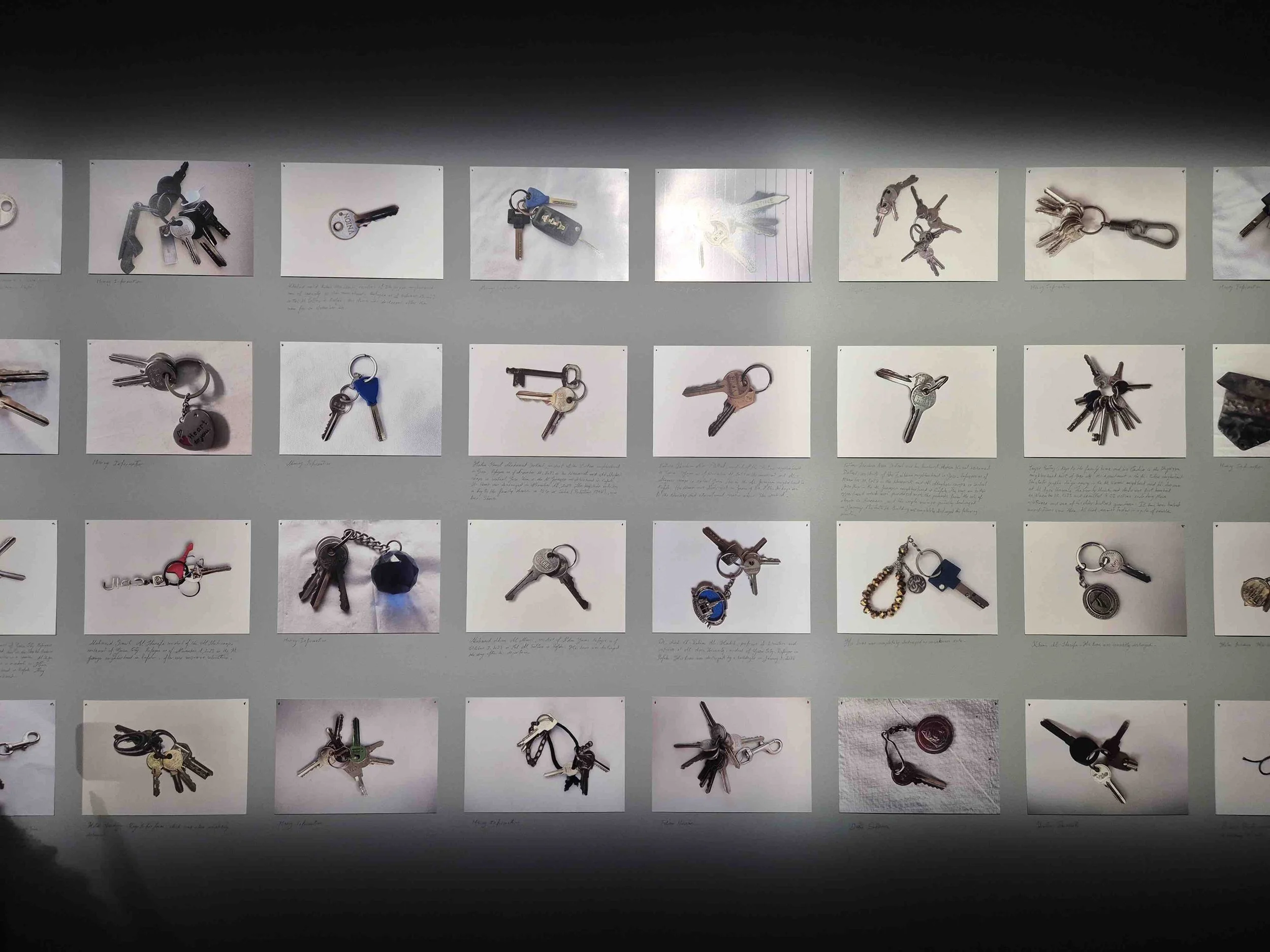 Display of various keychains and keys from abandoned houses in Palestine on a wall with written descriptions underneath each photograph.