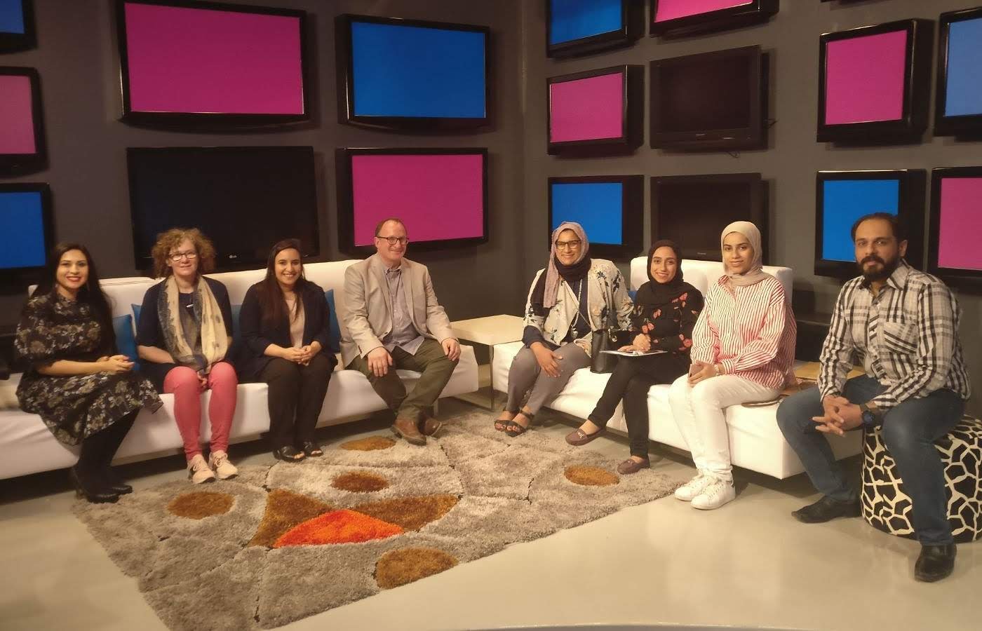 A group of nine diverse people sitting on white sofas in a room with multiple television screens on the wall, some turned off, some showing a pink and blue background.