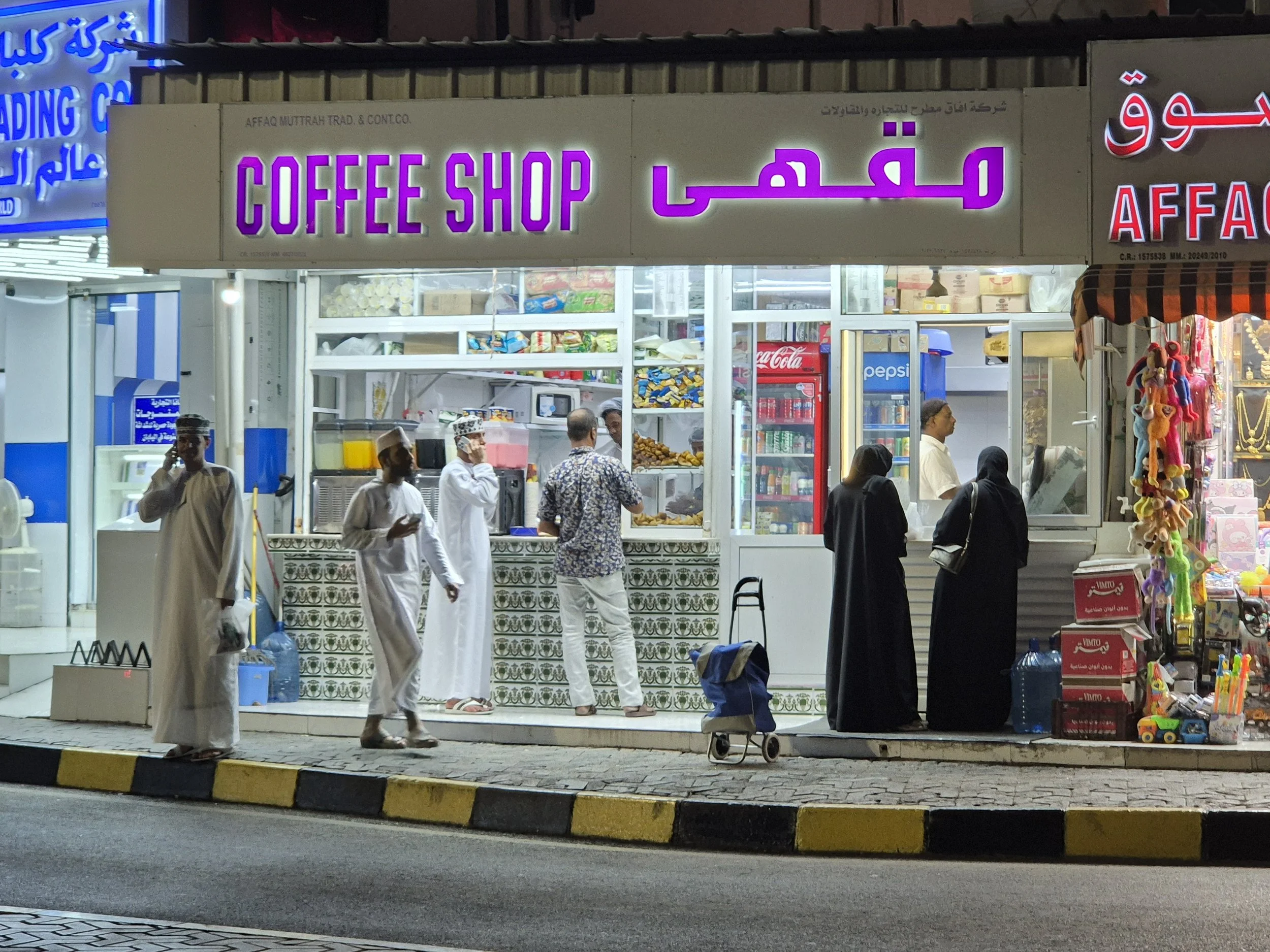 Night scene of a small coffee shop in Muttrah, Oman.  Several people are standing in line or waiting outside, some wearing traditional Middle Eastern attire and others in casual clothing. 