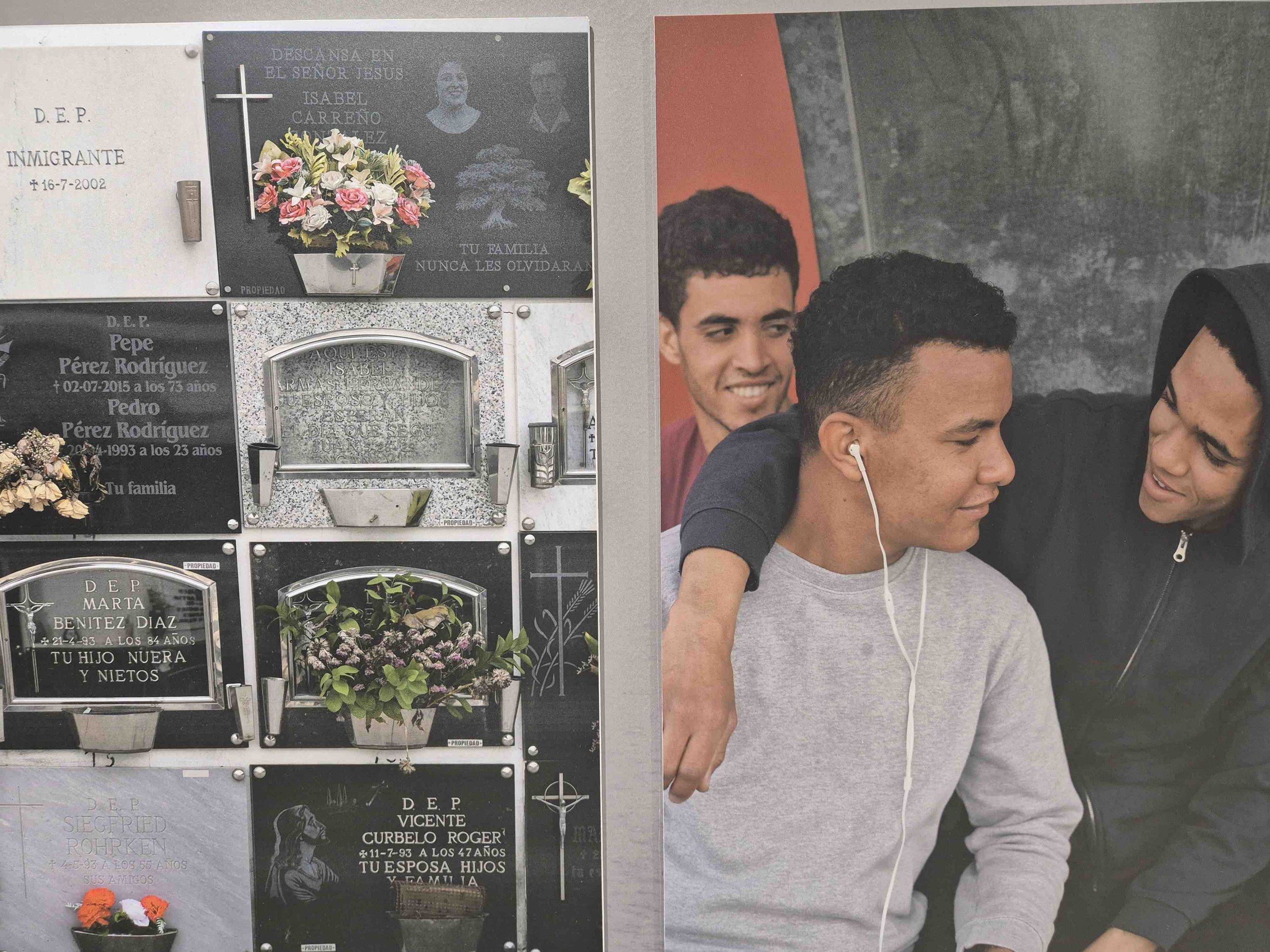 Young man in Sudan with earphones smiling while sitting between two friends, one of whom has his arm around him, in a cemetery with upright family tombstones attached to the wall.