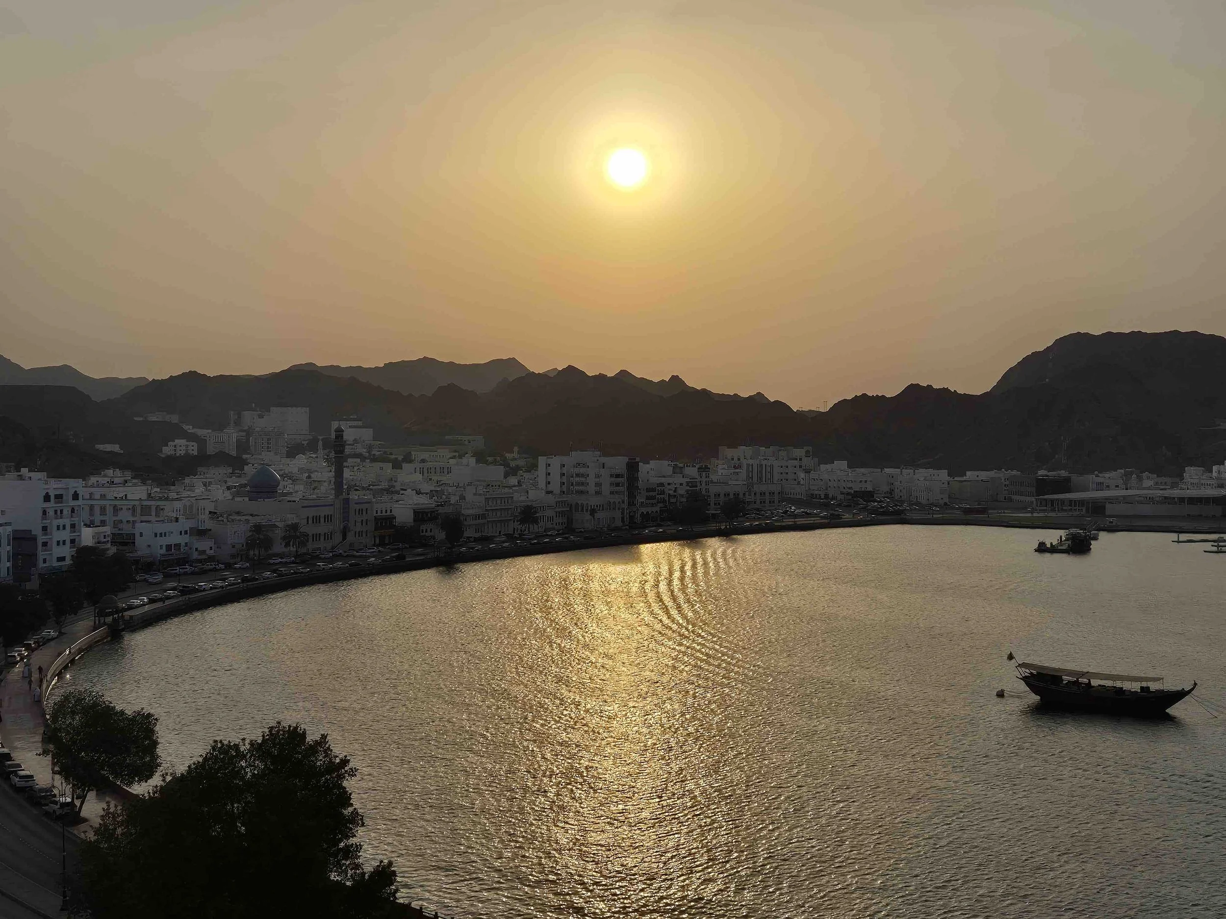Sunset over Muttrah, Oman, with a waterway, mountains in the background, and a boat on the water.