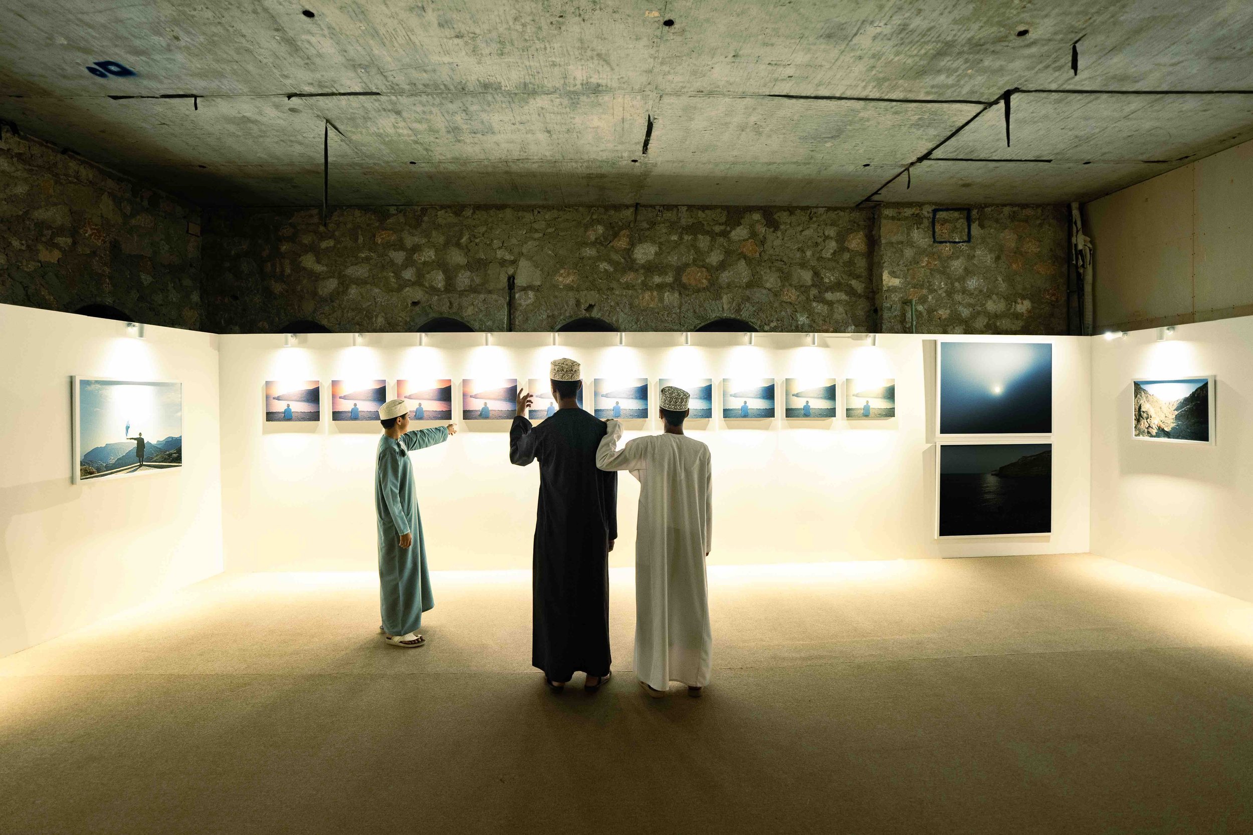Three men in traditional Middle Eastern attire, including robes and caps, viewing photographs in a temporary art gallery at the Raneen festival in Oman with white walls and a stone wall ceiling.