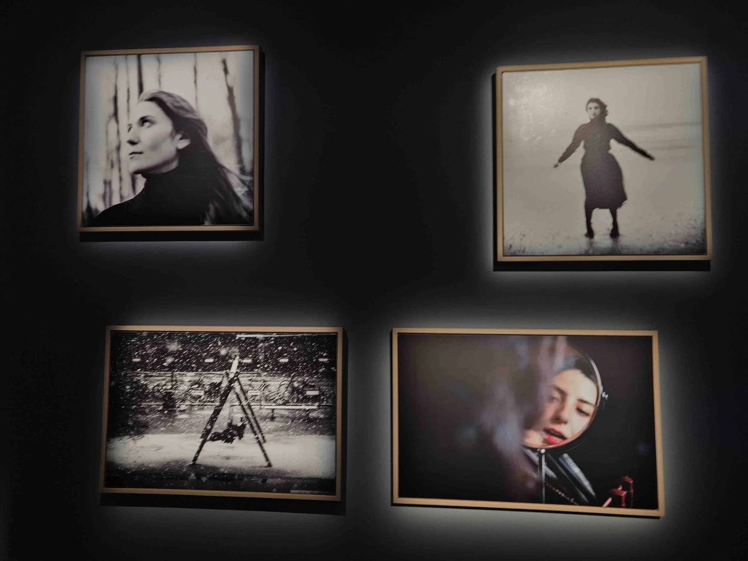 Four framed photographs displayed on a dark wall, including a black and white portrait of a woman, a person standing on a beach, a person on a swing in the snow, and a woman looking into a mirror.